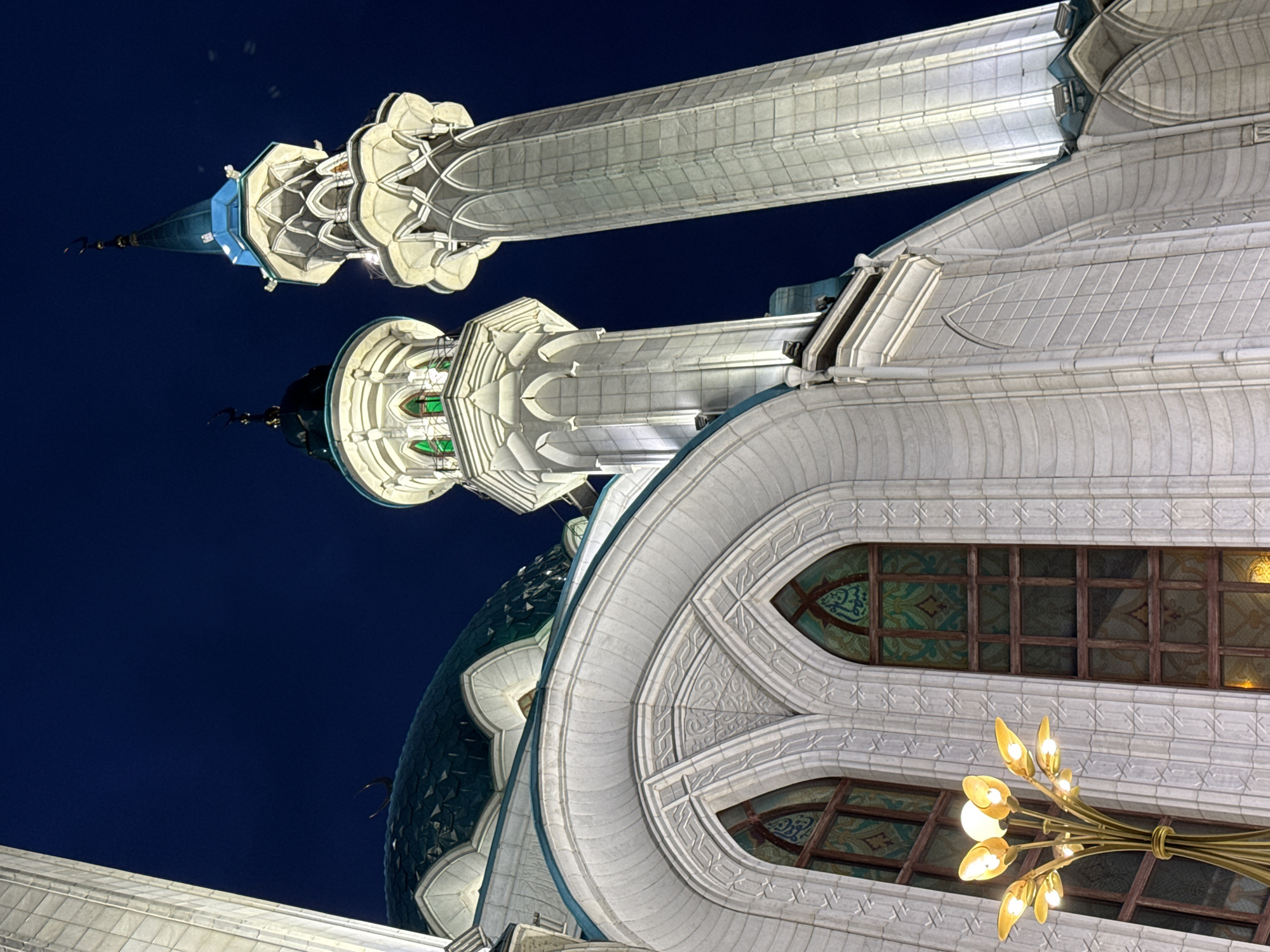 The image displays the majestic architecture of a mosque at night, set against a clear, deep blue sky. The scene is located in Kazan, Russia, likely depicting the Kul Sharif Mosque. The building is intricately detailed and brightly illuminated, highlighting its ornate features.
Two towering minarets dominate the upper portion of the frame. Both are constructed from light-colored stone with elaborate carvings and decorative balconies. One minaret is topped with a distinct blue, conical roof and a crescent finial, while the other features a darker, possibly green-hued, conical roof, also with a finial.
A large, textured dome, appearing in shades of teal or dark green with a scale-like pattern, is visible towards the left side of the structure. The main body of the mosque is built from light-colored stone, adorned with complex geometric patterns and arched window frames. The windows contain decorative stained glass panels, some of which subtly display green-toned Arabic calligraphy and traditional Islamic motifs.
In the bottom right foreground, a modern, golden-hued chandelier with multiple leaf-shaped light bulbs glows warmly, adding an interior element to the exterior view. The artificial lighting effectively emphasizes the architectural grandeur and delicate details of the mosque against the dark backdrop of the night sky, creating a serene and impressive atmosphere. The overall image captures the mosque's beauty and prominence after sunset.