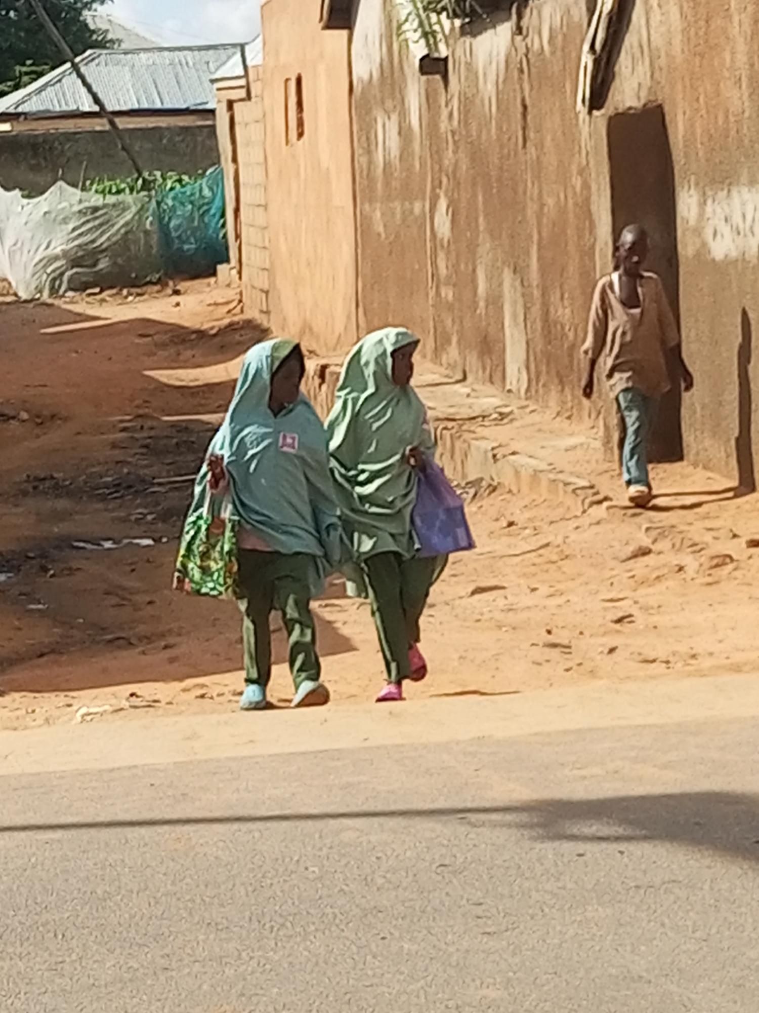 In the city of Tempure, Nigeria, two young females wearing light blue hijabs and matching green trousers walk side-by-side along a dirt road. The female on the left carries a multi-colored patterned tote bag, while the one on the right carries a solid purple shopping bag. Both are wearing matching colored hijabs, and the female on the right is wearing pink open-toed sandals. They are walking on the edge of a paved road, in front of a row of mud-brick buildings. The buildings have corrugated metal roofs and arched doorways. Further back, there is a large building with a metal roof, partially covered by a white tarp. The sky is clear and blue. A third person, an adult male wearing a brown shirt and blue jeans, walks in the background away from the camera.