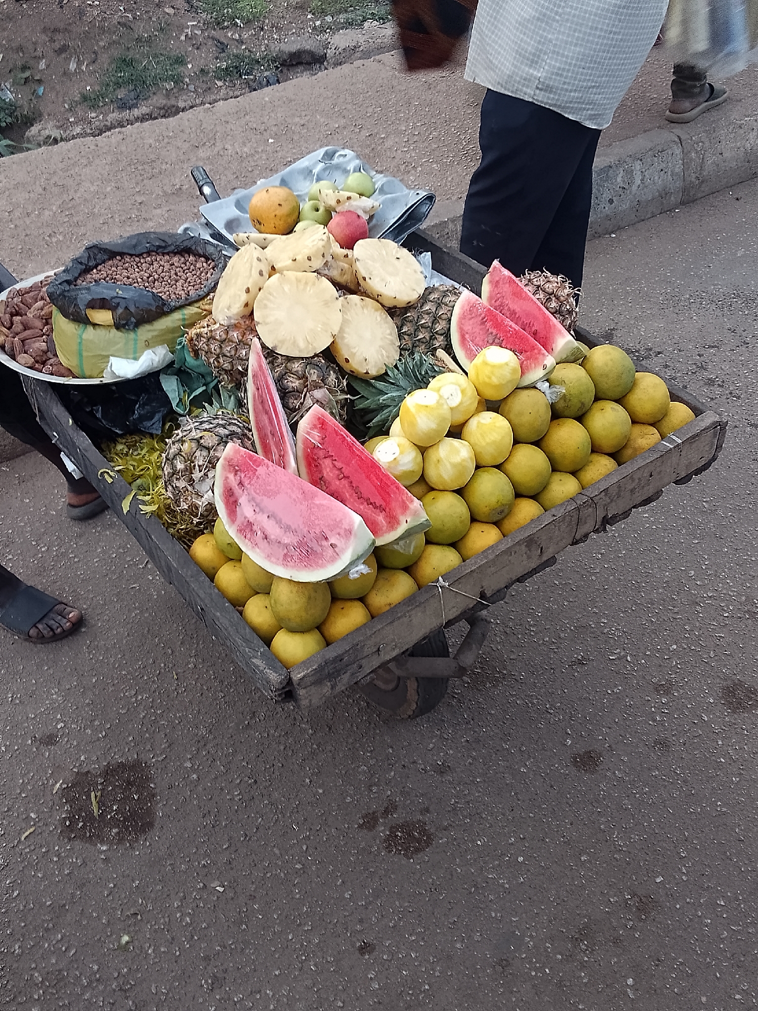 A street vendor in Bukuru, Nigeria, stands by a wooden cart filled with a variety of fruits and nuts. The cart, positioned on an asphalt road, contains peeled oranges, sliced pineapples, watermelon slices, whole pineapples, dates, groundnuts in a plastic bag, and assorted fruits in a styrofoam tray. The vendor wears black pants and a light-colored, checkered shirt. The background features a concrete barrier and sparse vegetation. The road surface is textured with dark patches and small debris.