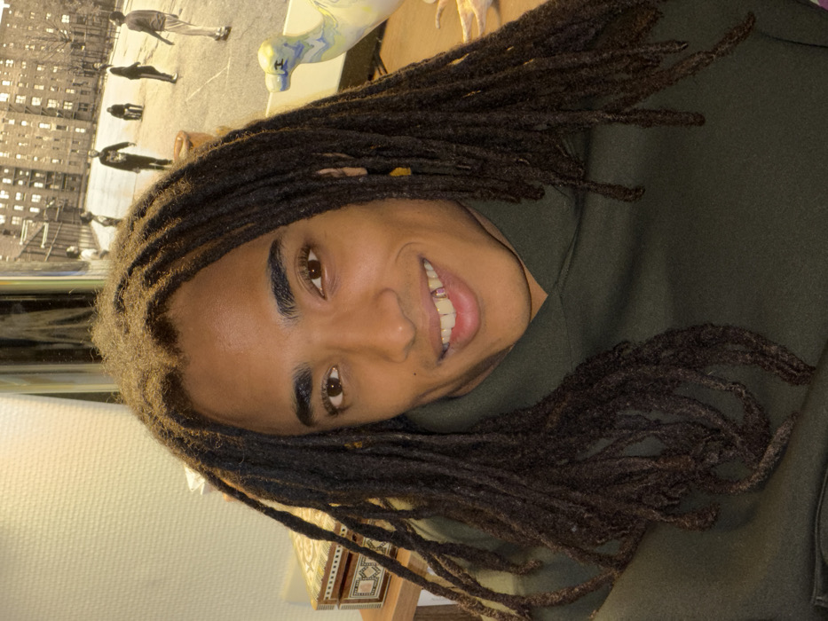 The image displays a young individual with dark skin and long, dark brown dreadlocks, oriented on their side as the image is rotated 90 degrees clockwise. The person is smiling broadly, looking directly towards the viewer with warm brown eyes. Their facial features include well-defined, dark eyebrows, a visible upper row of teeth, and a slight glint on what appears to be a dental adornment or filling on one of their upper front teeth. They are wearing a dark green hooded sweatshirt.
The background provides a glimpse into an indoor setting. To the left side of the person's head (which would be the top if the image were upright), there is a large, framed print or poster. This print features a black and white or sepia-toned scene depicting several people walking across what looks like a street or open area, with multi-story buildings visible in the background, suggesting an urban or residential environment. The figures in the print are distinct, with one appearing closer to the foreground and others further back.
Above the person's head and extending to the right, there is a light-colored, possibly ceramic or painted, abstract decorative object or sculpture that vaguely resembles an animal figure, possibly a dog or horse, with blue and yellow swirls of color on its surface. Further to the bottom right of the image, below the person's shoulder and arm, a corner of a decorative wooden box with intricate patterns and possibly mother-of-pearl or similar inlay is visible, resting on a surface. The lighting appears soft and warm, illuminating the person's face. No text is visible in the image.