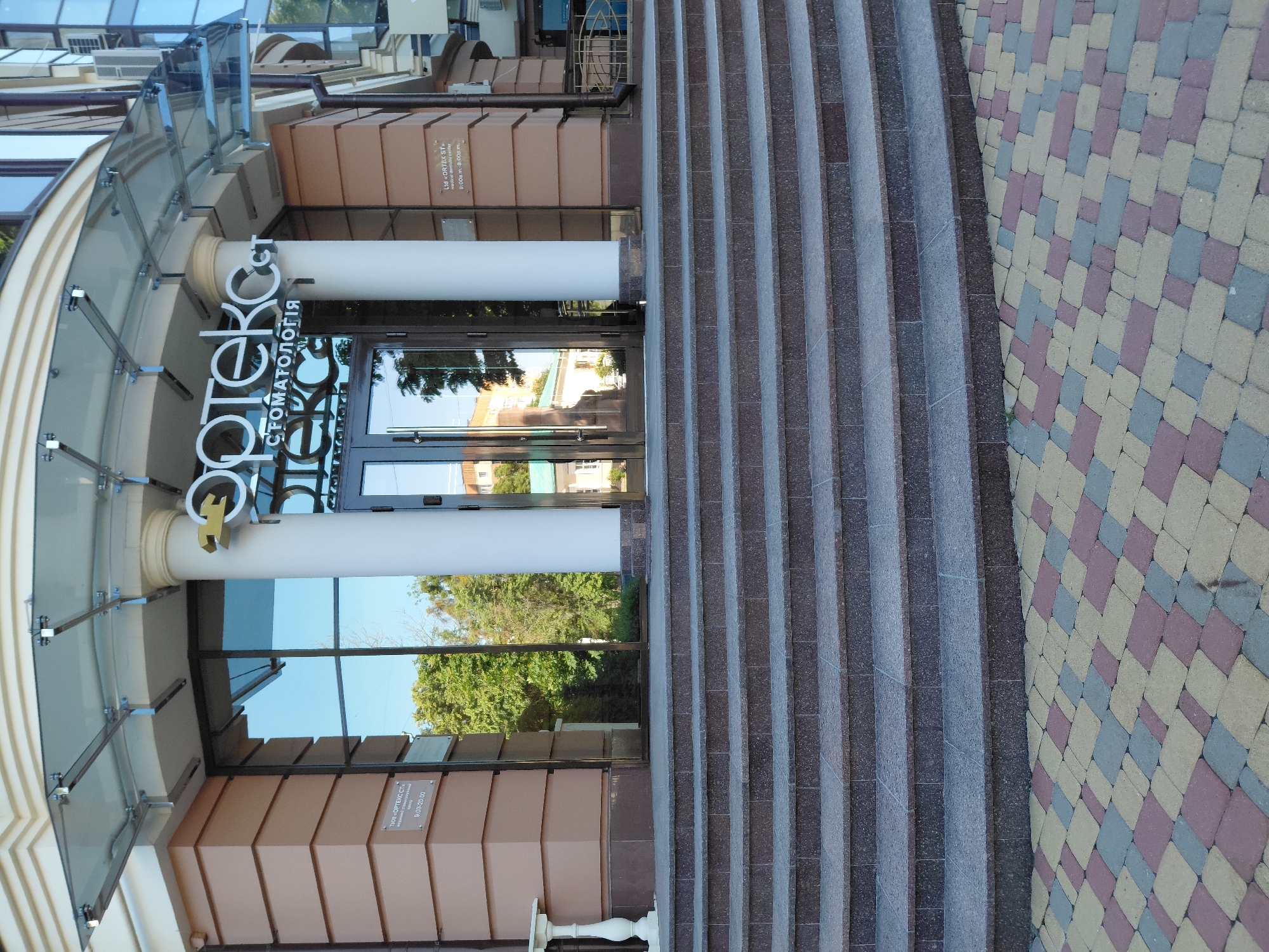 A horizontally oriented photograph depicts the modern entrance of a medical dentistry center, featuring a prominent set of steps, glass doors, and signage. The scene is devoid of people, focusing solely on the building's facade and its immediate surroundings.
The main subject is the building's entrance, accessible via a wide set of steps crafted from grey and dark speckled stone. Above the glass entrance doors, a curved glass canopy extends outwards, supported by metal fixtures. Two large white columns flank the doors, adding to the architectural design. The large glass panels of the doors and the upper windows reflect a clear blue sky and lush green trees, indicating a sunny daytime setting.
To the right of the steps, a patterned paved area is visible, constructed from rectangular blocks in shades of beige, light blue, and reddish-brown, arranged in a diagonal grid.
Prominently displayed above the entrance is a large white sign with the name "Ортекс" (Ortex) and "СТОМАТОЛОГІЯ" (Dentistry) in Cyrillic letters. A smaller rectangular plaque is affixed to the building's facade, providing details in both Ukrainian and English: "ТОВ «ОРТЕКС СТ» / Ltd «ORTEX ST»", "медичний стоматологічний центр / medical dentistry center", and the operating hours "9:00-20:00 / 9:00a.m.-8:00p.m." The overall impression is that of a professional and well-maintained commercial establishment.
