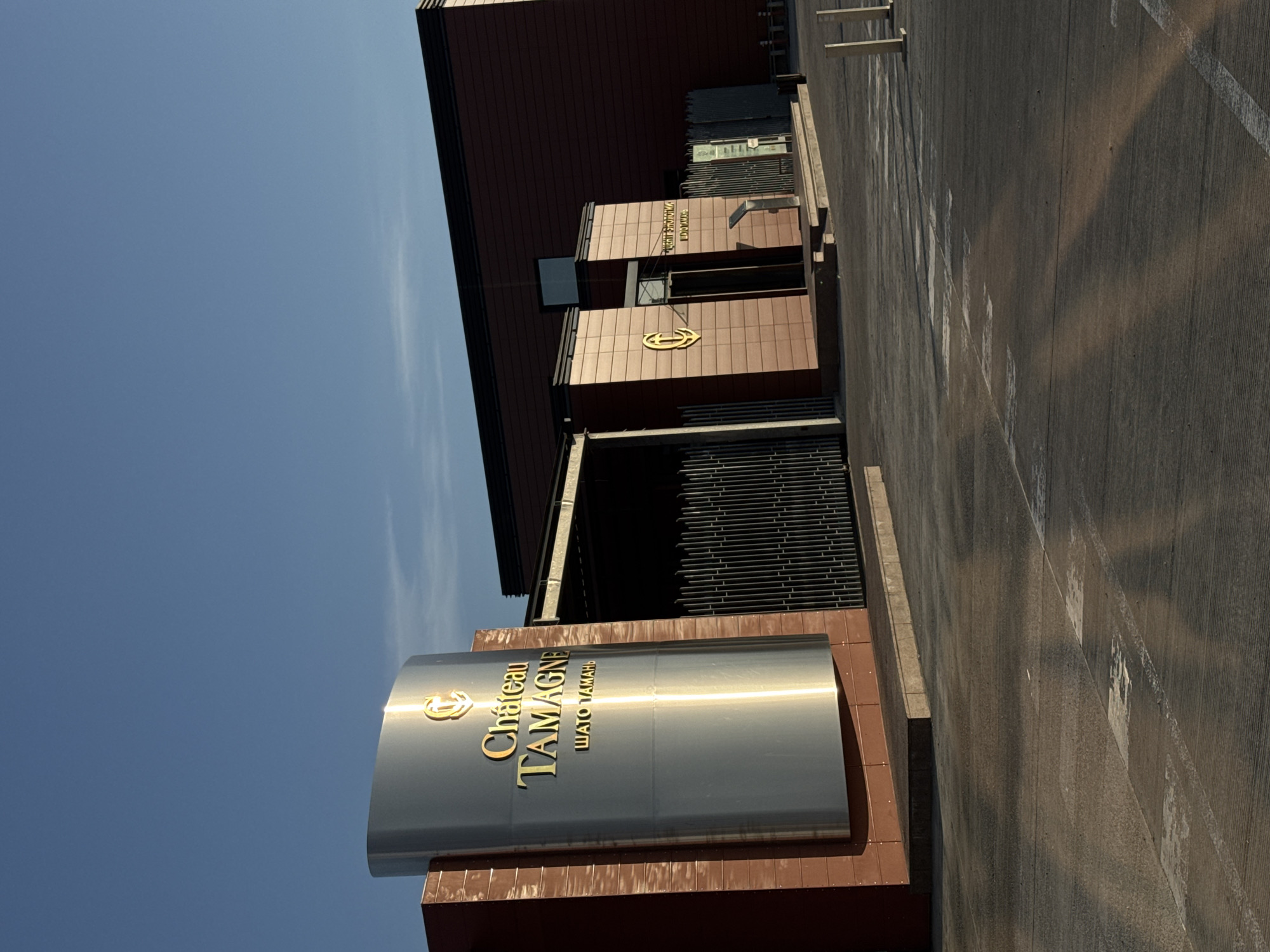 The image depicts the exterior of Chateau Tamagne in Taman, Russia. The architectural style appears modern, with a combination of dark brown cladding and metal accents. A prominent curved sign bearing the Chateau Tamagne name in gold lettering is positioned in front of the building. The surroundings include a paved parking area and a clear blue sky. There are no visible people in the frame.