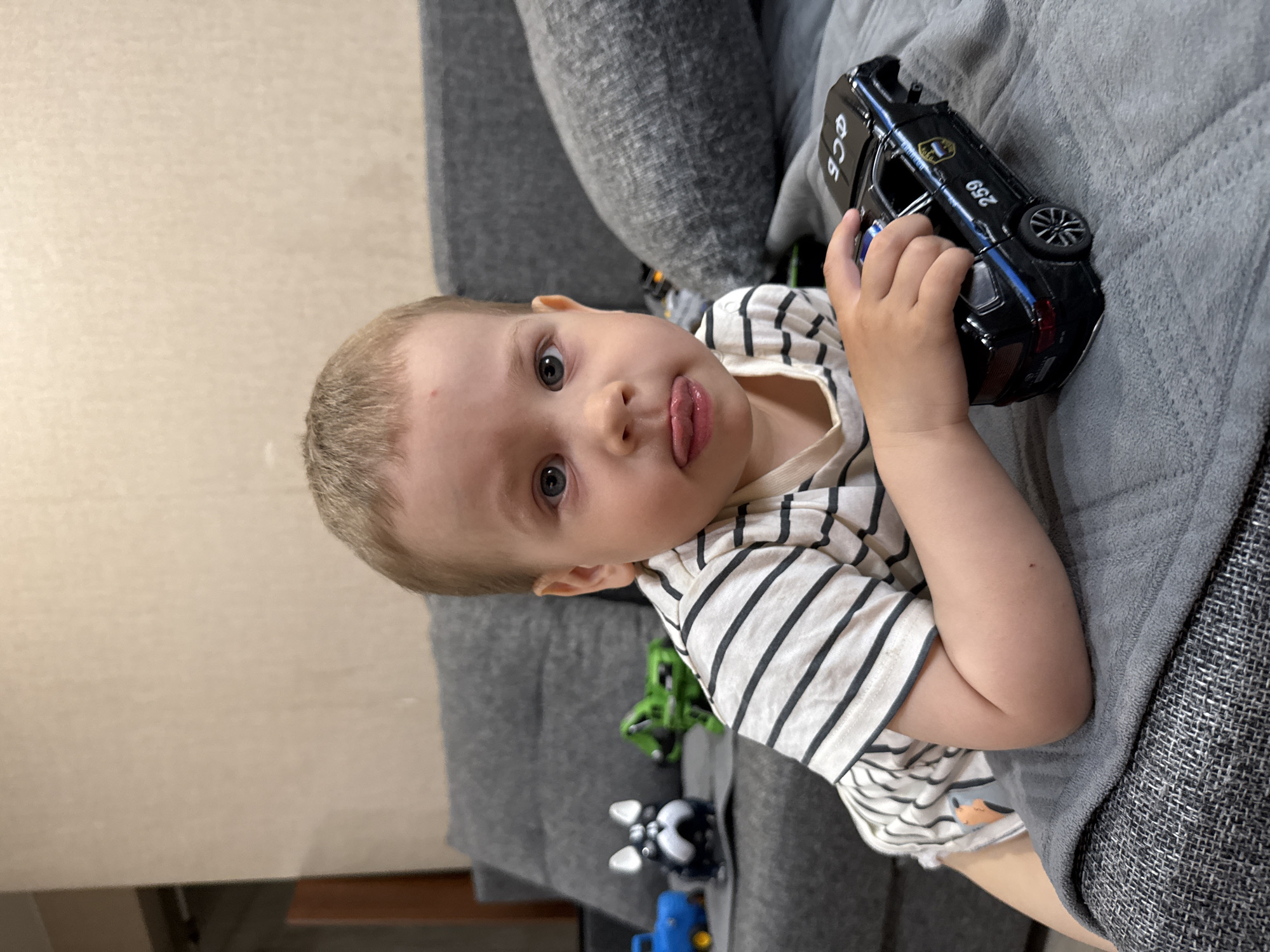 A young child with fair skin and light-colored hair is lying on a gray couch, sticking their tongue out towards the camera. The child is wearing a striped shirt with white and black lines. The child is holding a black toy police car with the number "259" on it. There are other toy vehicles visible on the couch as well. The setting appears to be indoors, with a beige wall visible in the background. The location is the city of Unknown, Unknown.