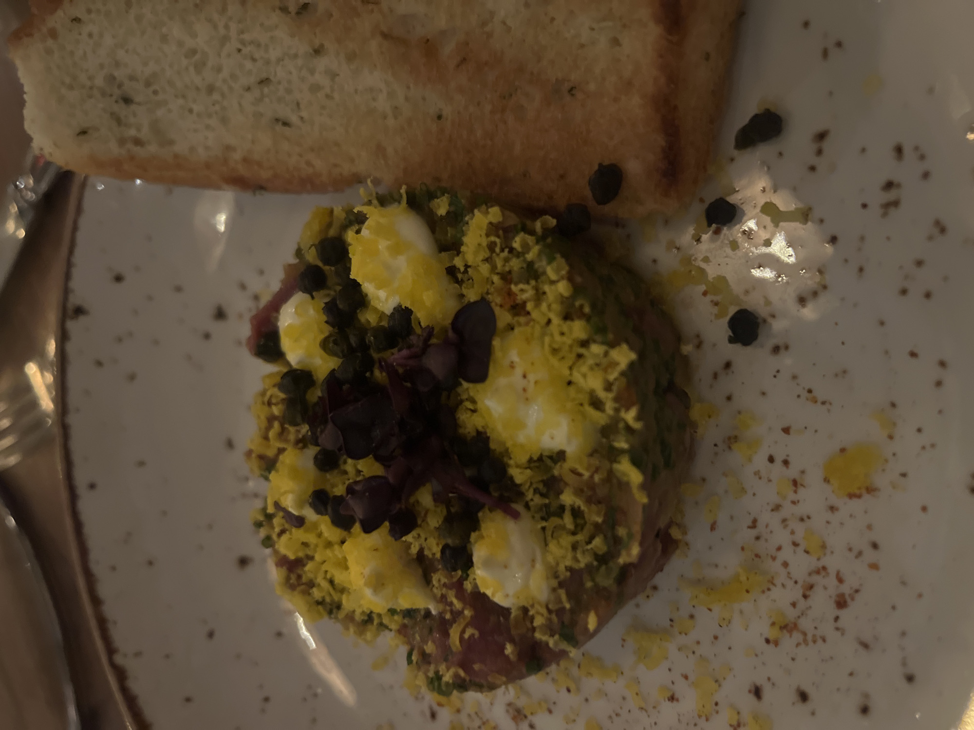 The image depicts a dish of food presented on a white plate. A piece of toasted bread is positioned at the top of the frame. Below the bread is a mound of finely chopped ingredients, including what appear to be meat, herbs, and eggs. A few dark-colored capers and small purple sprouts garnish the dish. The plate has some scattered crumbs and drizzles of oil. The image appears to be taken indoors, likely in a restaurant setting. The location, Berlin, Germany, is not identifiable from the image itself.