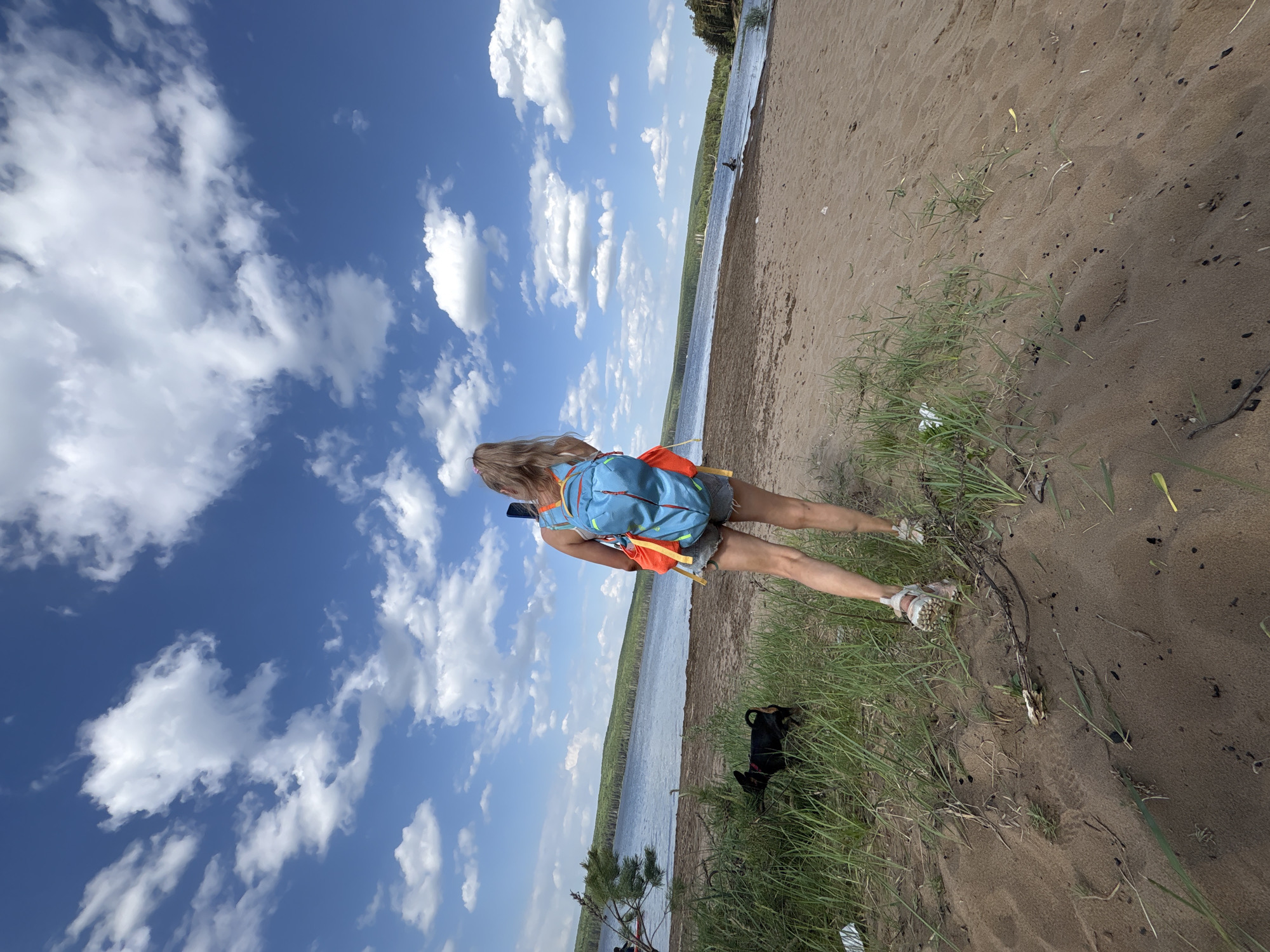A woman with long blonde hair is standing on a sandy beach near a body of water, possibly the Kama River. She is wearing a backpack and appears to be taking a picture with her phone. A small black dog is nearby in the grass. The sky is blue with scattered white clouds. The image was taken in the city of Chaikovsky, Russia.