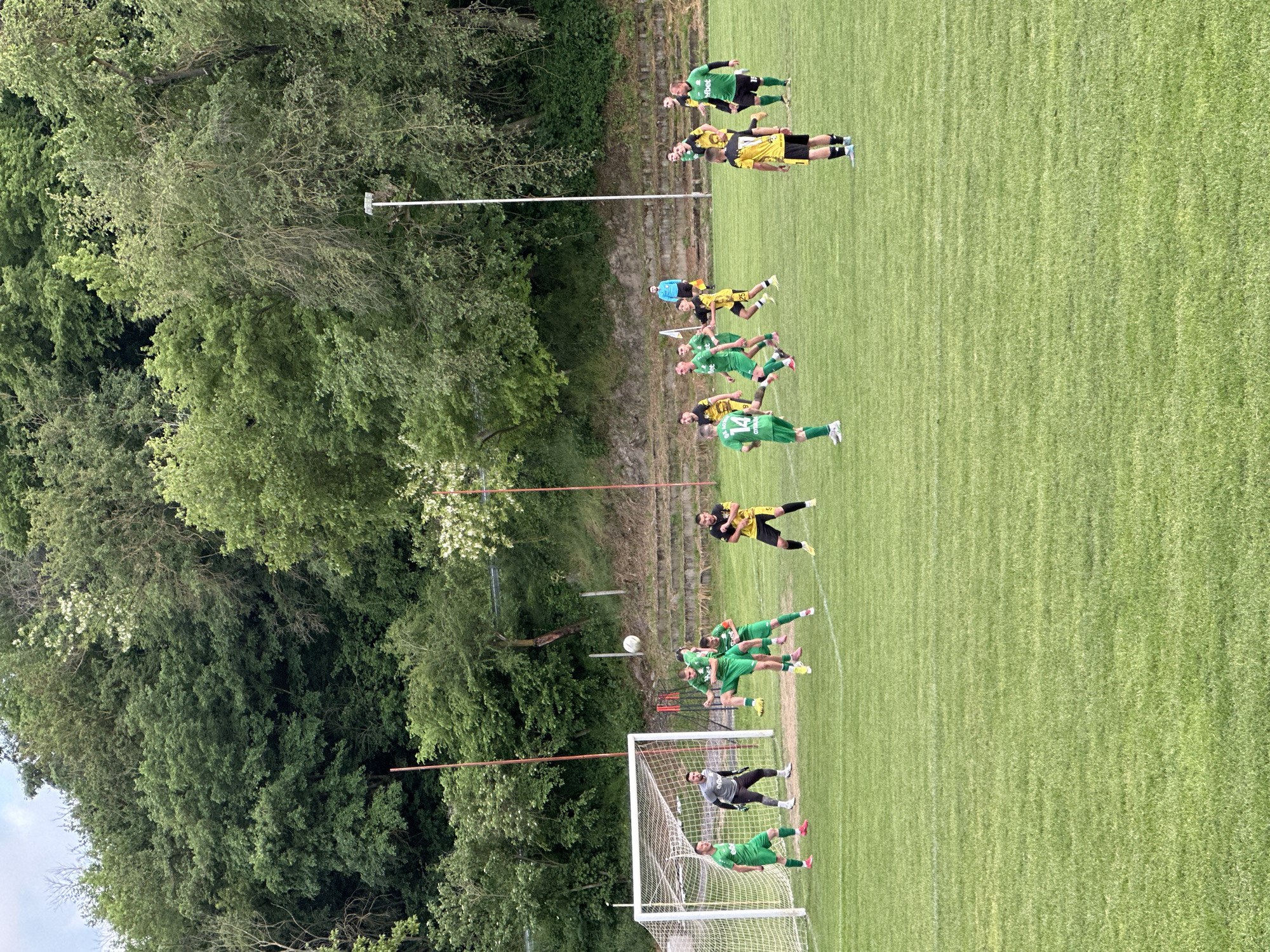 The image depicts a soccer game in progress, likely a free kick situation, on a grassy field in Debelets, Bulgaria. The scene includes a soccer goal with a goalkeeper positioned inside. A group of players in green jerseys are lined up in front of the goal, presumably forming a defensive wall. Another player in a yellow jersey stands near the ball, preparing to take the kick. A small group of spectators can be seen on a slight elevation behind the line of players. The background features a dense area of trees. The field markings are visible, and the overall lighting suggests it is daytime. The exact location in Debelets is not discernible from the image.