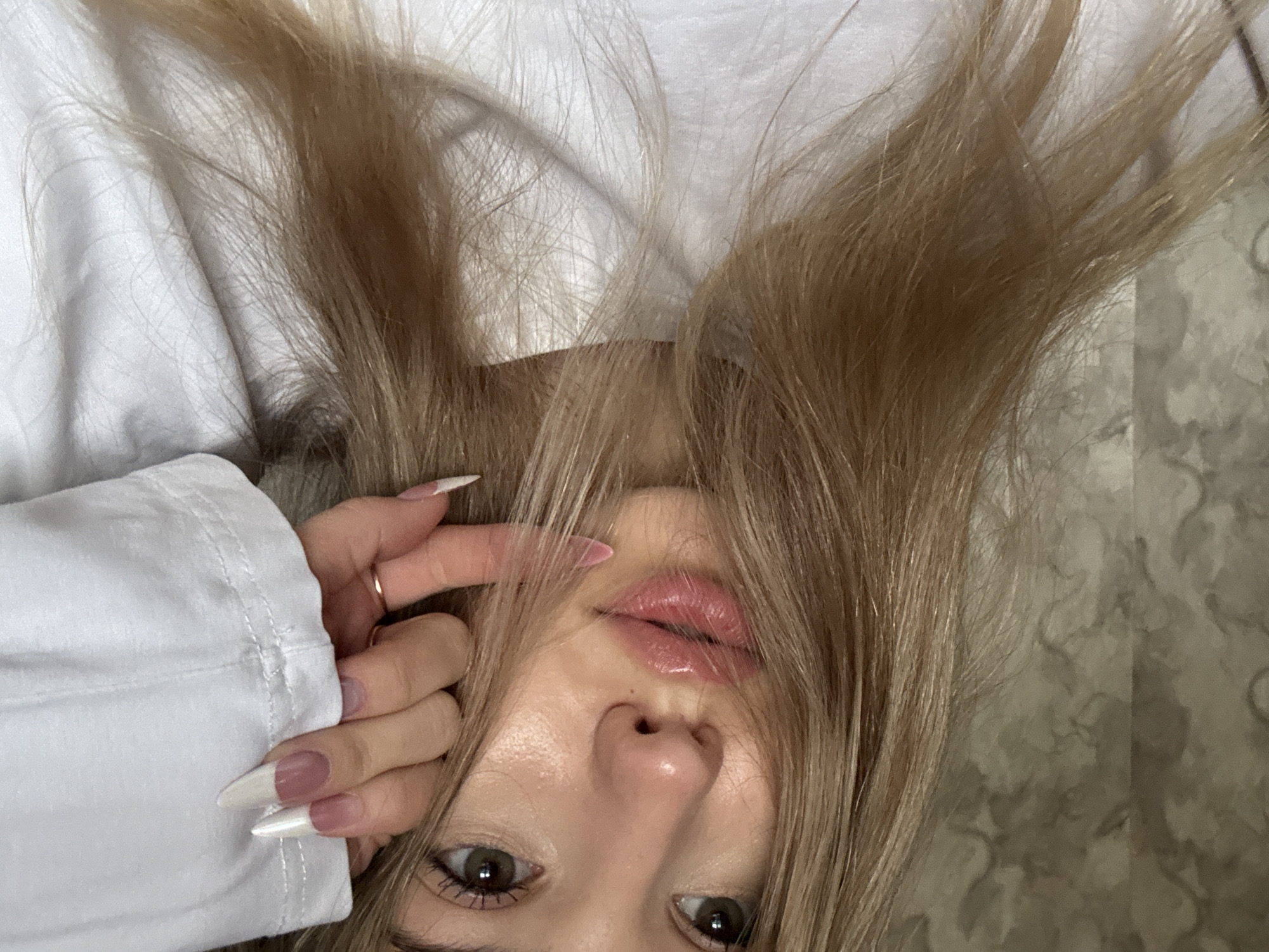 A close-up, upside-down selfie shows a woman with long, light blonde hair partially obscuring her face. Her face features prominent lips with a glossy sheen, a slightly upturned nose, and light eyes with dark pupils. She wears a white long-sleeved shirt, and a gold ring is visible on her finger. Her fingernails are long and painted with a pale pink base and white tips. The background consists of a white crumpled fabric and a beige surface with a light pattern. The image was taken in an indoor environment and does not contain information to identify the city of Noviy, Russia.