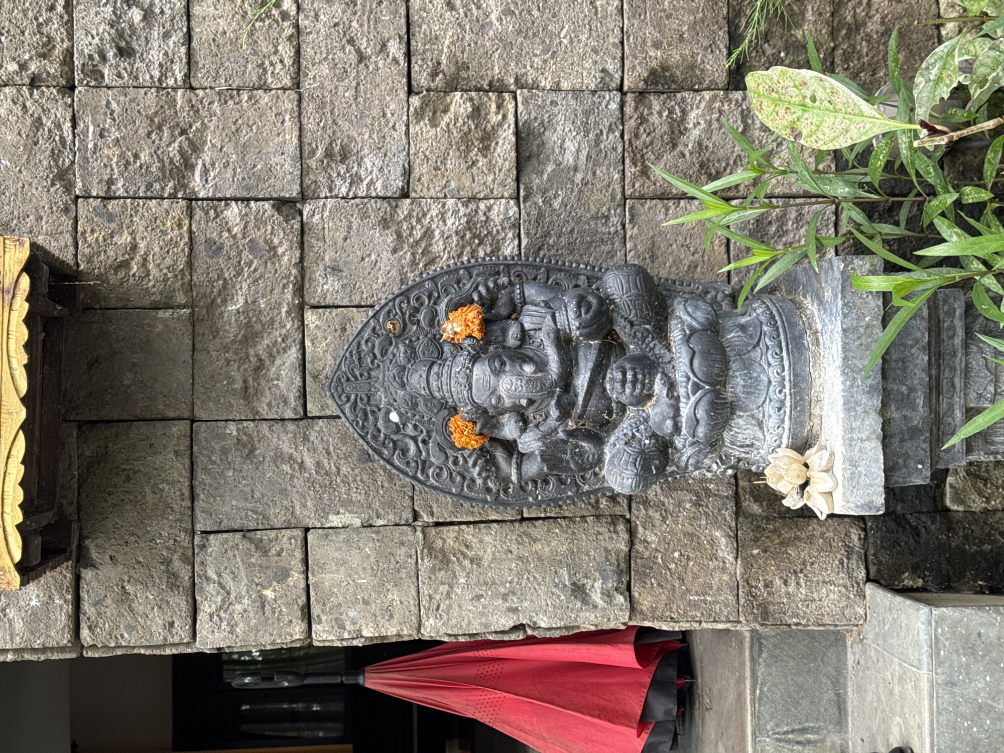 The image displays a dark, intricately carved stone statue, likely depicting the Hindu deity Ganesha, resting horizontally on a textured surface of grey, rectangular stone pavers. The statue features an elephant head, multiple arms, and ornate detailing. Several small, orange, marigold-like flowers are placed on the statue's head and body, serving as offerings. A cluster of small, light-colored or white flowers is also visible near the right side of the statue's base.
The background consists of more grey stone pavers and lush green foliage from various plants in the upper right. A partial view of a folded, red umbrella with black details is visible in the lower left corner, lying on a darker surface beneath the pavers. To the far left, a portion of a golden, carved wooden object adds another decorative element to the scene.
The scene is brightly lit by natural daylight, indicating daytime. The clear lighting emphasizes the textures of the stone, the statue's carvings, and the fresh greenery. The presence of the Ganesha statue and flower offerings strongly suggests a cultural or religious context, indicative of a sacred space or decorative display often found in places like Kuta Selatan, Indonesia. No discernible text is visible within the frame.