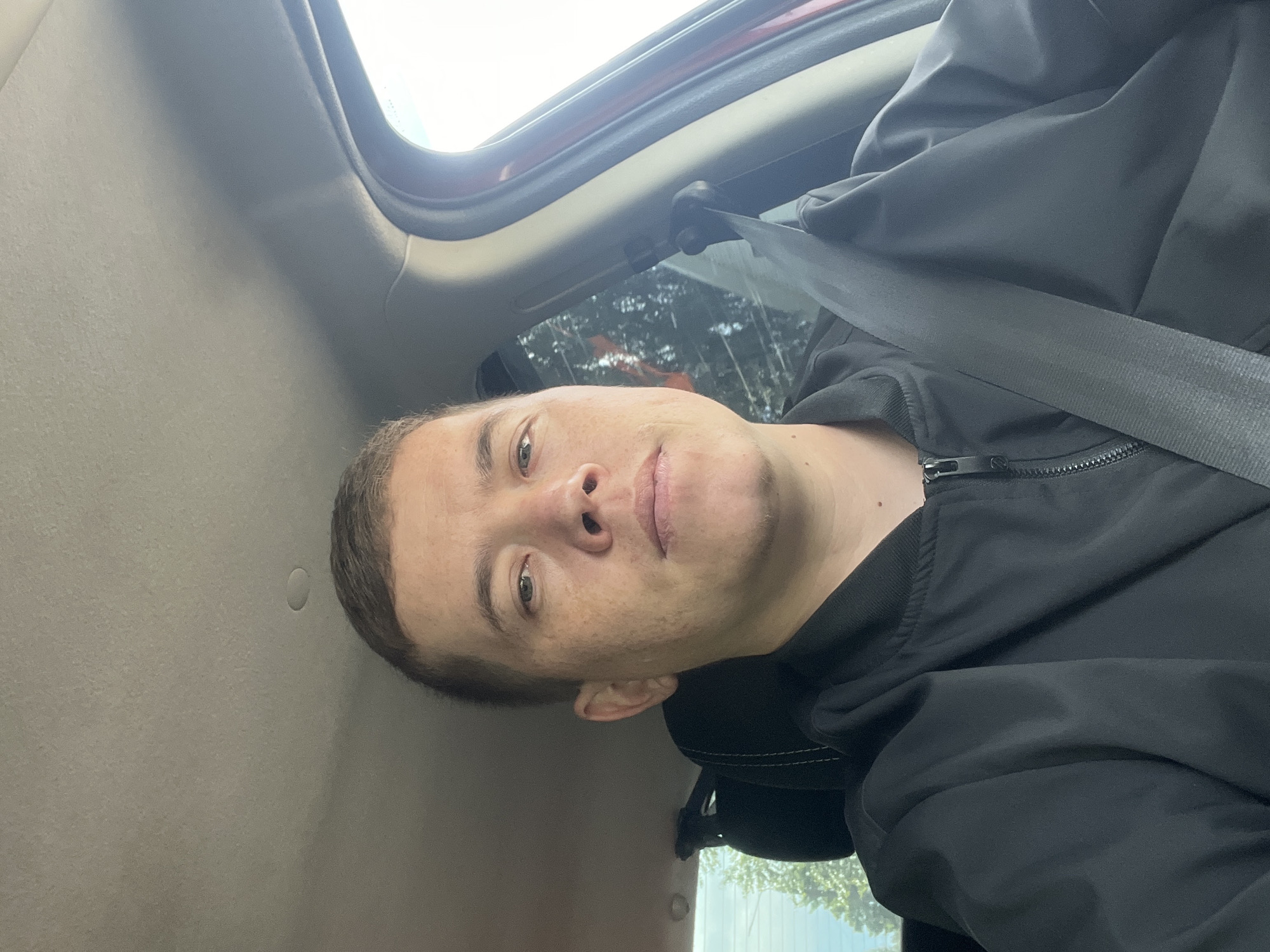 A male individual, appearing to be in his 30s or 40s, with short brown hair and a light complexion featuring scattered freckles, is depicted in the foreground. He is wearing a black, zippered bomber-style jacket and a dark-colored seatbelt across his chest and left shoulder. His expression is neutral as he looks directly into the camera. The setting is the interior of a vehicle, visible by a light-colored fabric headliner, a dark headrest behind the subject, and parts of the car's door frame and A-pillar to his right. Through the car windows, natural daylight is visible, along with green foliage, indicating an outdoor daytime scene. The specific location is the city of Izhevsk, Russia.