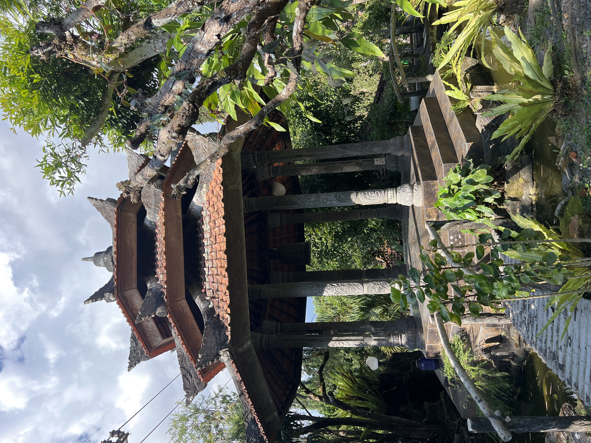The image depicts an outdoor scene featuring a multi-tiered, octagonal pavilion with a red-tiled roof, characteristic of Balinese architecture. The pavilion is supported by ornate, carved stone pillars. A large bell hangs inside the pavilion. The structure is elevated and accessed by stone steps. A rustic wooden bridge with a simple railing spans a narrow body of water that appears to be a pond or stream, with a small waterfall or rocky edge visible. Lush green vegetation, including ferns and trees, surrounds the pavilion and water feature, suggesting a garden or natural setting. The sky above is partly cloudy with a mix of blue sky and white clouds. There is a blue plastic container and a white lamp post near the pavilion. A power line is visible in the upper left corner. The scene is identified as being in the city of Pupuan, Indonesia. There are no people visible in the image.