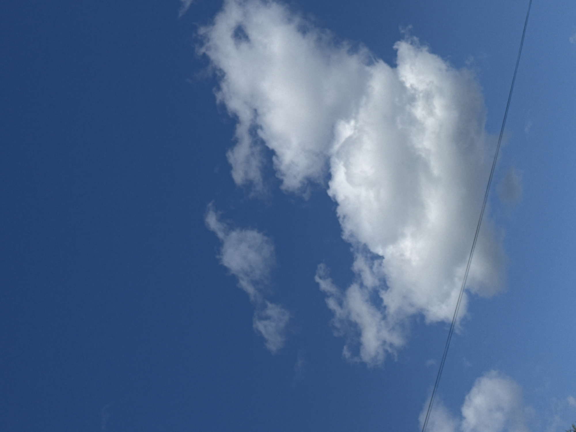 A vertical, medium shot captures a bright blue sky with scattered white clouds. Two thin, dark power lines stretch diagonally across the lower portion of the frame, from the bottom left to the upper right. The clouds are fluffy and illuminated by the sun, with one large cloud formation dominating the center of the image. A few smaller, wispy clouds are present in the upper left and lower right corners. The scene appears to be an outdoor shot with no people or specific structures visible besides the power lines, suggesting an open area like a park or a residential street with overhead utilities. There is no information about the specific location within Cheboksary, Russia.