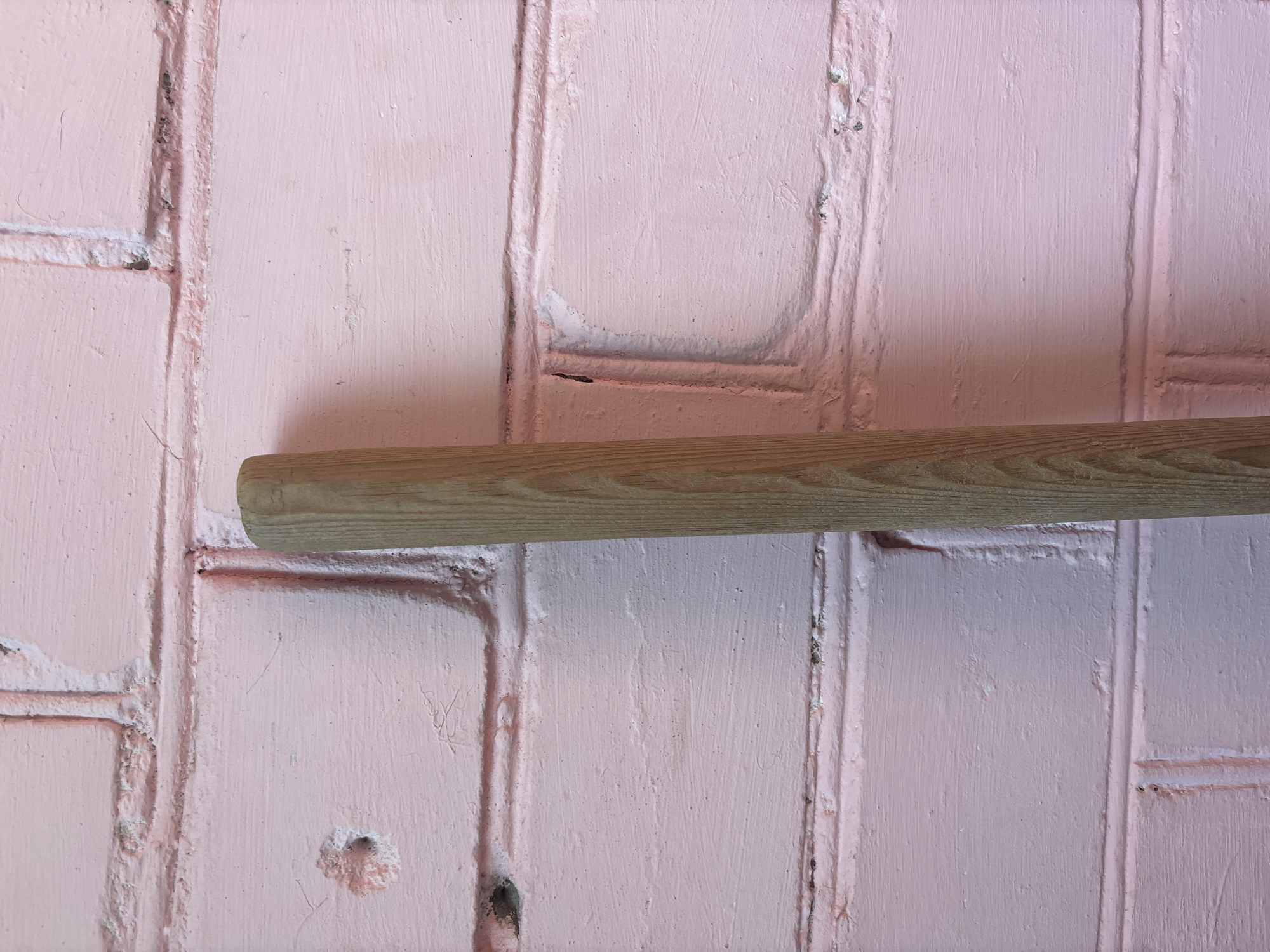 A close-up, eye-level, vertical shot shows the side of a wooden handle of a tool, possibly a broom or mop, set against a pink brick wall. The handle occupies the right side of the frame, with its rough wooden texture visible. The brick wall, painted pink, fills the background. The bricks are laid horizontally, with visible mortar lines and some imperfections on the painted surface, such as small holes and rough patches. The lighting appears to be natural, casting subtle shadows and highlighting the textures of both the wood and the brick. The scene is static and appears to be outdoors or in a space with similar lighting conditions.