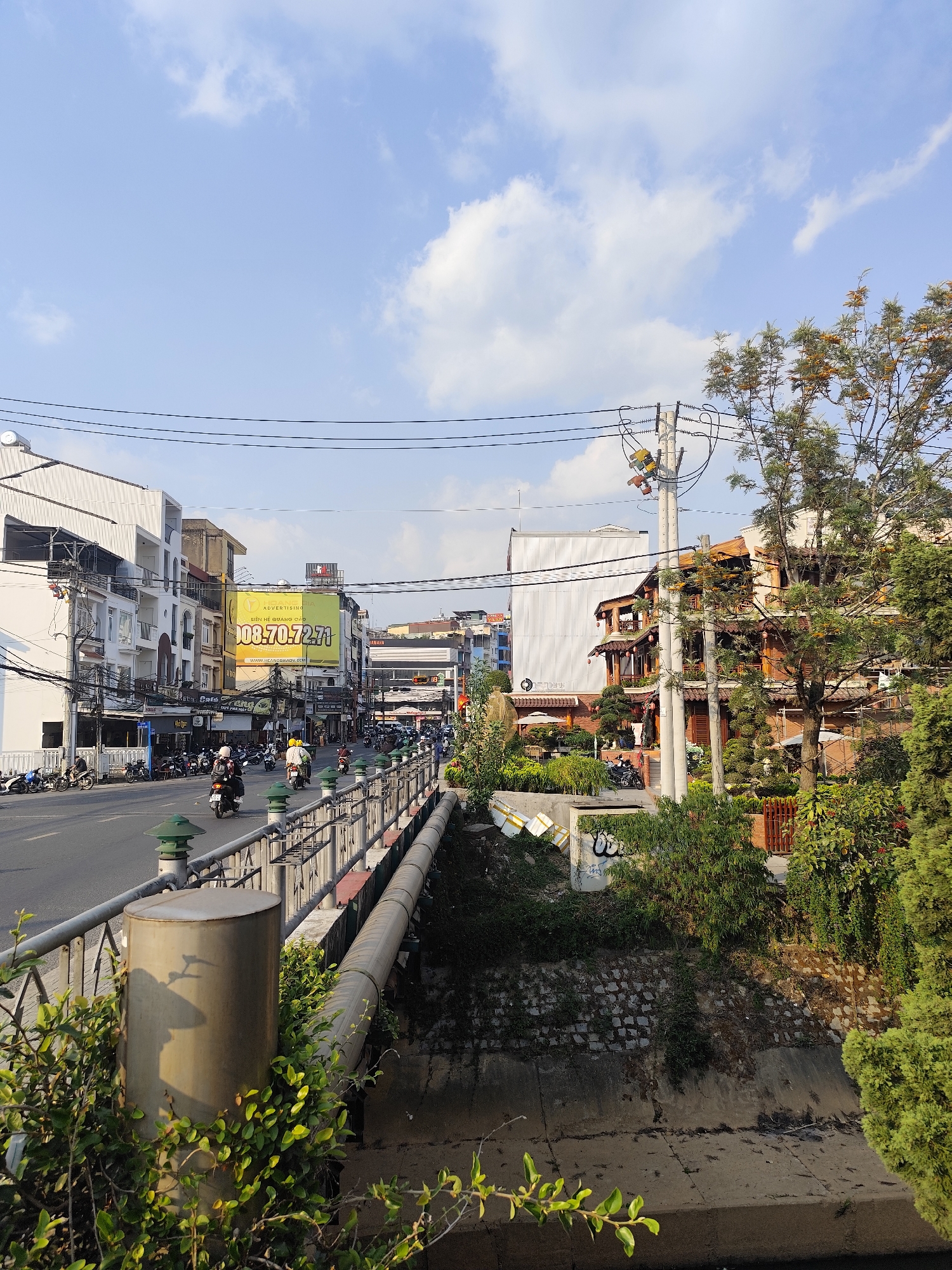 The image presents a daytime urban street scene in Da Lat, Vietnam, under a blue sky with scattered white clouds. The view is from an elevated vantage point, looking down a street.
In the immediate foreground, a metal pedestrian railing with green-capped decorative posts runs parallel to a paved road. Below this railing, a large, light-colored pipe is attached horizontally. Further below, a concrete-lined canal or drainage ditch extends from foreground to background. The canal's far bank is composed of a stone wall, partially overgrown with vegetation and marked with white graffiti. Discarded white rectangular objects are visible along the top edge of the canal wall. A gray cylindrical post is partially obscured by green foliage in the lower left.
On the left, a two-lane asphalt road carries multiple motorcycles traveling away from the viewer. Riders are visible wearing helmets, one notably in a yellow helmet and jacket. The left side of the street is lined with multi-story buildings, primarily commercial or mixed-use. A prominent yellow billboard on one building displays "HÒA NGHĨA QUẢNG CÁO" and a contact number "0908.70.72.71." Numerous motorcycles are parked along the sidewalk in front of these buildings.
On the right side of the scene, a dark utility pole with transformers and numerous overhead electrical wires stands prominently. Adjacent to it, a large tree with green and some yellowish-orange foliage provides shade. Beyond the dense greenery and closer to the right, a multi-story building with traditional wooden architectural elements, balconies, and red lanterns is visible. Its lower section features brickwork, and an outdoor seating area with white umbrellas is nestled among landscaped plants.
The background shows additional multi-story city buildings extending down the street on both sides.