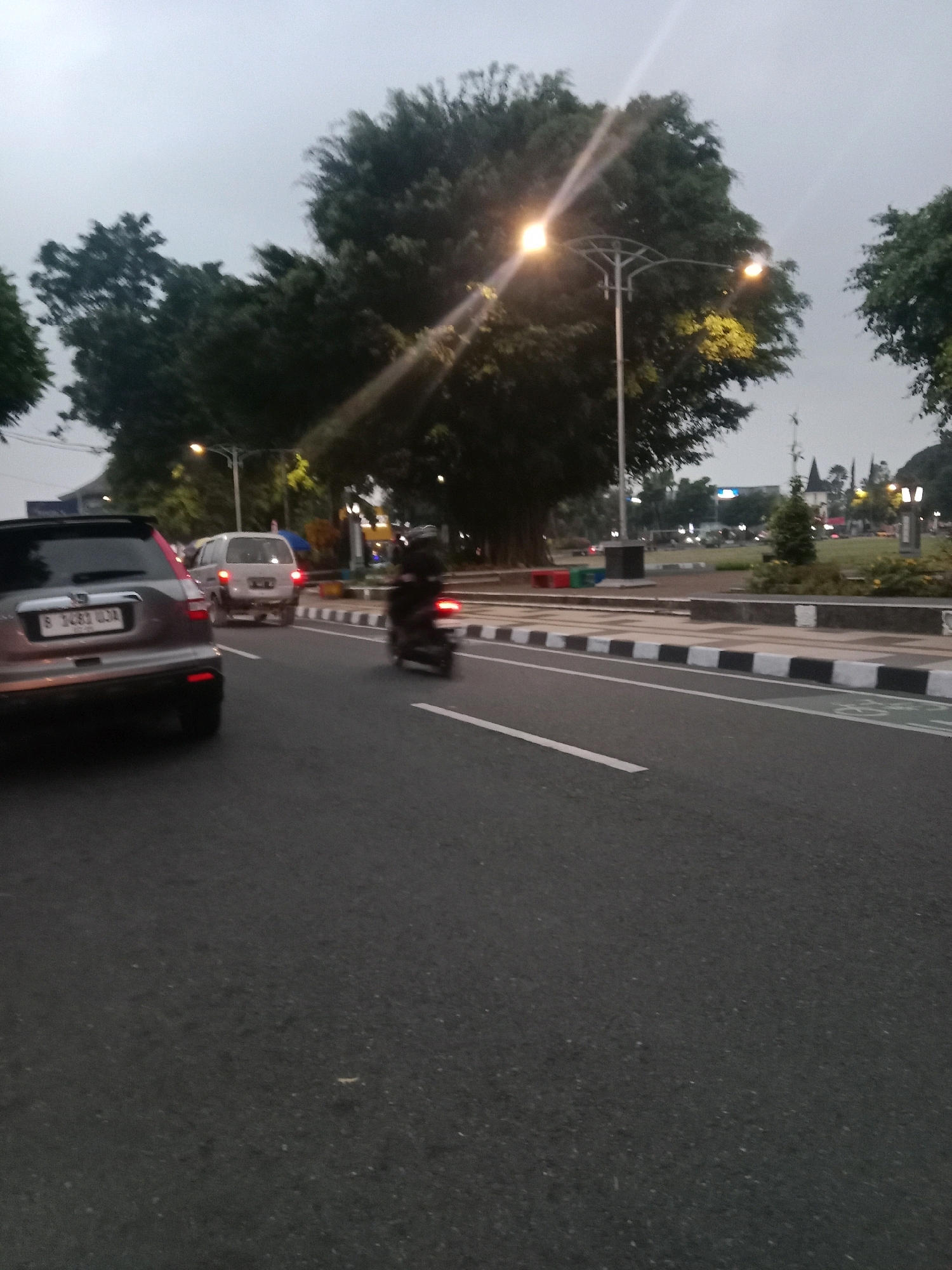 The image depicts a street scene in the city of Wonosobo, Indonesia, likely during overcast conditions or early morning/late evening due to the diffused light and illuminated street lamps. In the foreground, a grey SUV is visible from the rear, parked or moving slowly in the left lane. Its license plate reads "B 1483 UJA". To the right of the SUV, a white van is also visible in the adjacent lane, with its rear facing the camera. Further ahead, a motorcycle rider is moving in the right lane, with their form blurred due to motion. The road is paved asphalt with distinct white lines marking the lanes and a white and black striped curb along the right side. Behind the road, a grassy area with trees and street lamps is visible. In the background, there are more trees and some buildings and structures, including a church-like spire. The overall impression is that of a typical urban or semi-urban street in Indonesia.