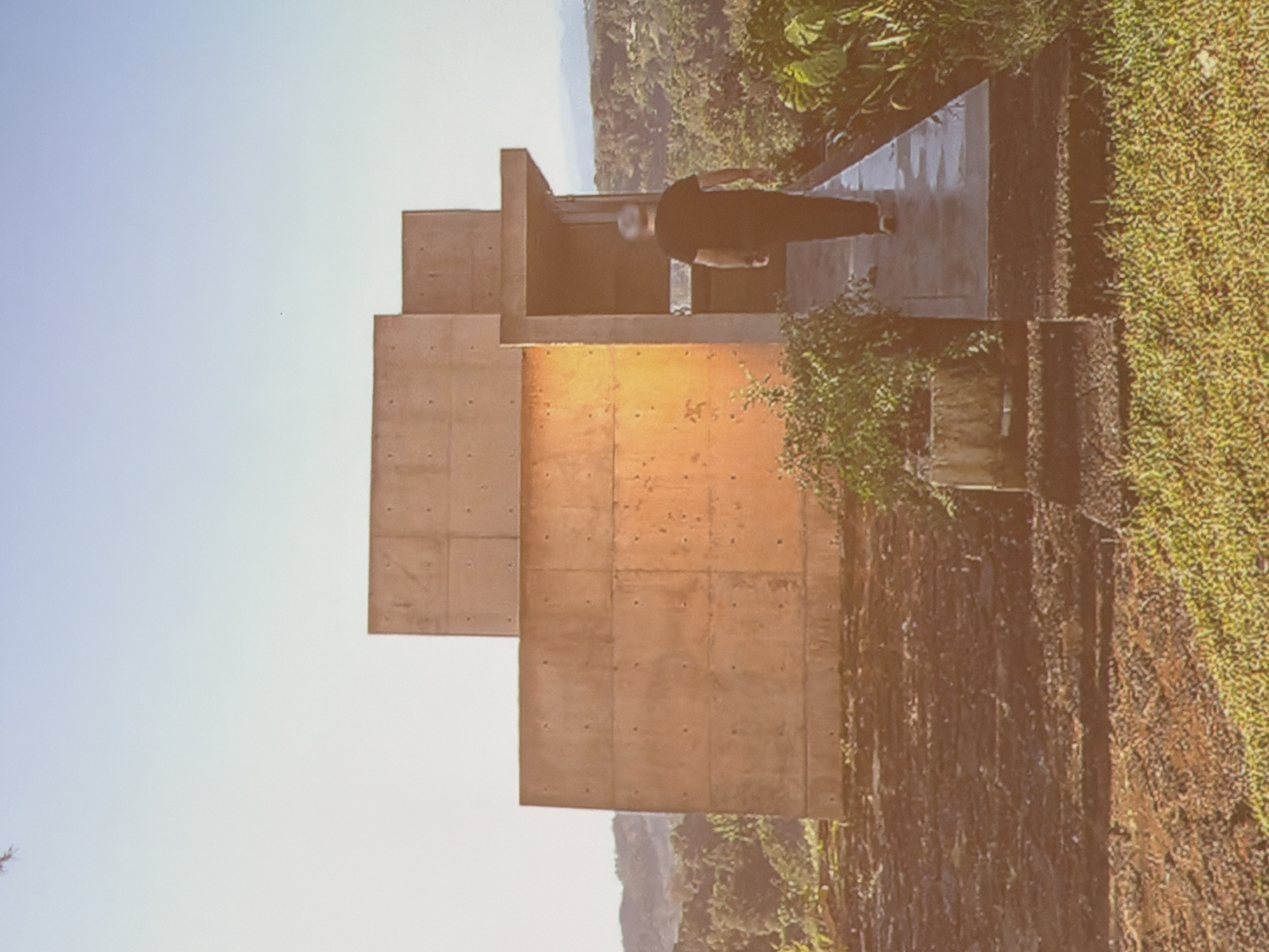 A building made of concrete is situated on top of a mountain. A person is standing on top of the concrete building. There is foliage on the right side. In the distance, there are additional mountain peaks visible. The sky is blue. It is located in the city of Unknown, Unknown.
