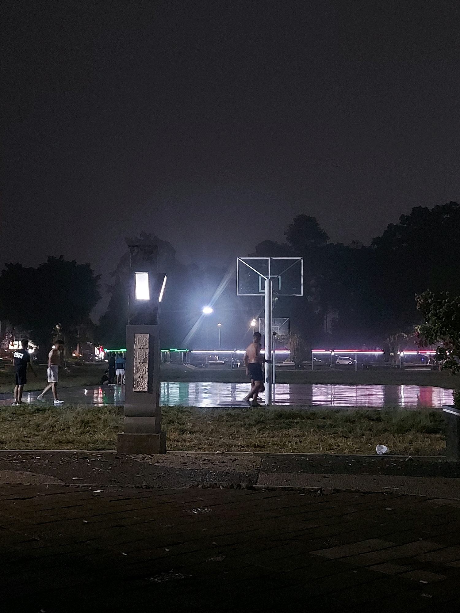 A basketball court at night in Wonosobo, Indonesia, is dimly lit by artificial lights. The court appears wet, reflecting the lights and creating a shimmering effect. Several people are present, some of whom are playing basketball. One individual, shirtless and wearing shorts, is walking across the court. Another person, wearing a black t-shirt with the number "1982" on it, stands with another individual wearing white shorts. A tall, illuminated structure with decorative carvings stands near the court. In the background, trees are visible, and more lights suggest other areas of activity or infrastructure. The overall atmosphere is dark and somewhat hazy, characteristic of a damp night.