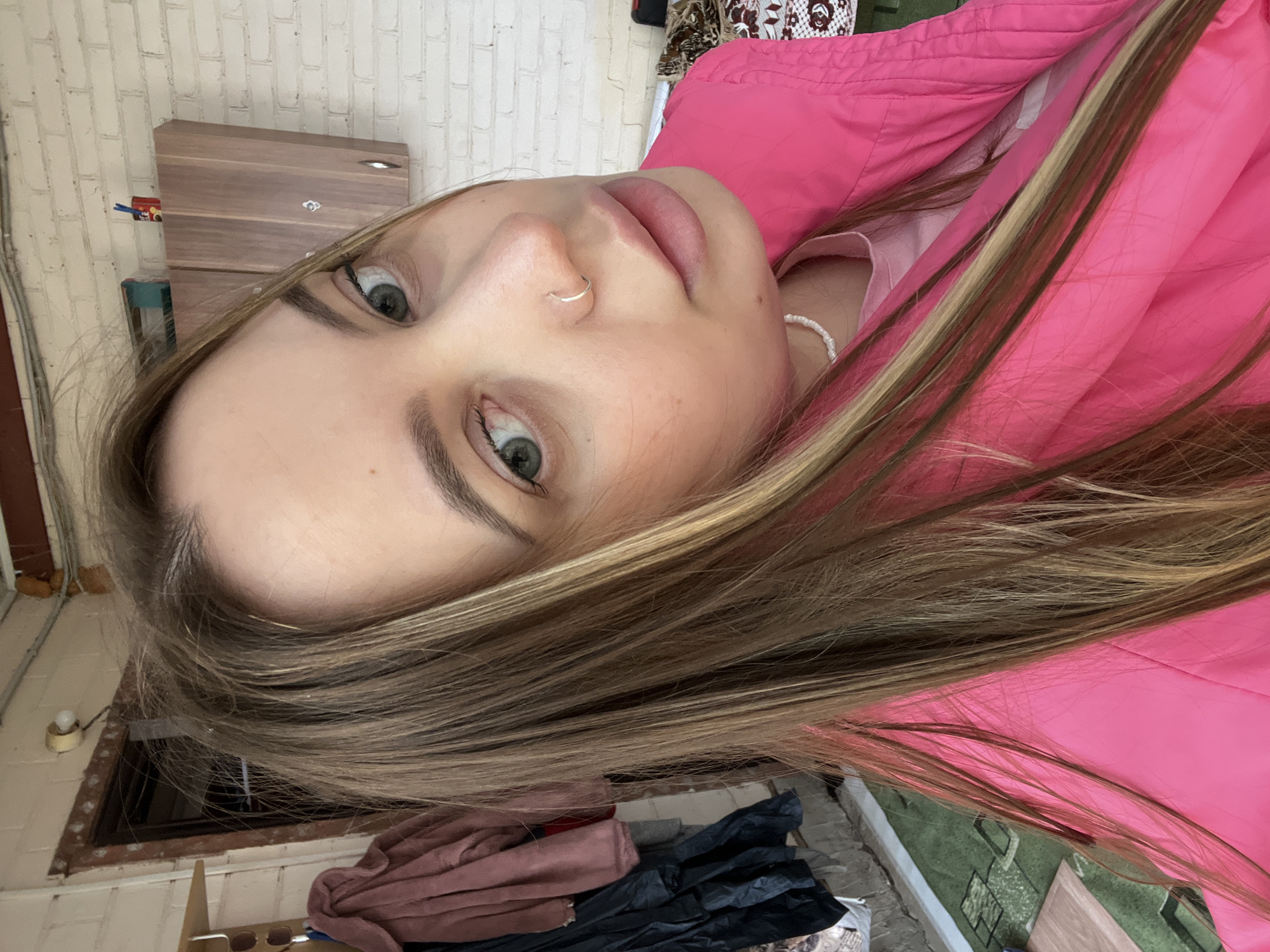 A close-up, vertically oriented photograph shows a woman with fair skin, light brown hair with blonde highlights, and green eyes. She has a silver nose ring in her left nostril. She is wearing a pink jacket and a white beaded necklace is visible. The background includes a brick wall, a wooden cabinet, and an outdoor area with miscellaneous items including clothes, plants and other debris. The location is suggested as Novopokrovskaya, Russia.