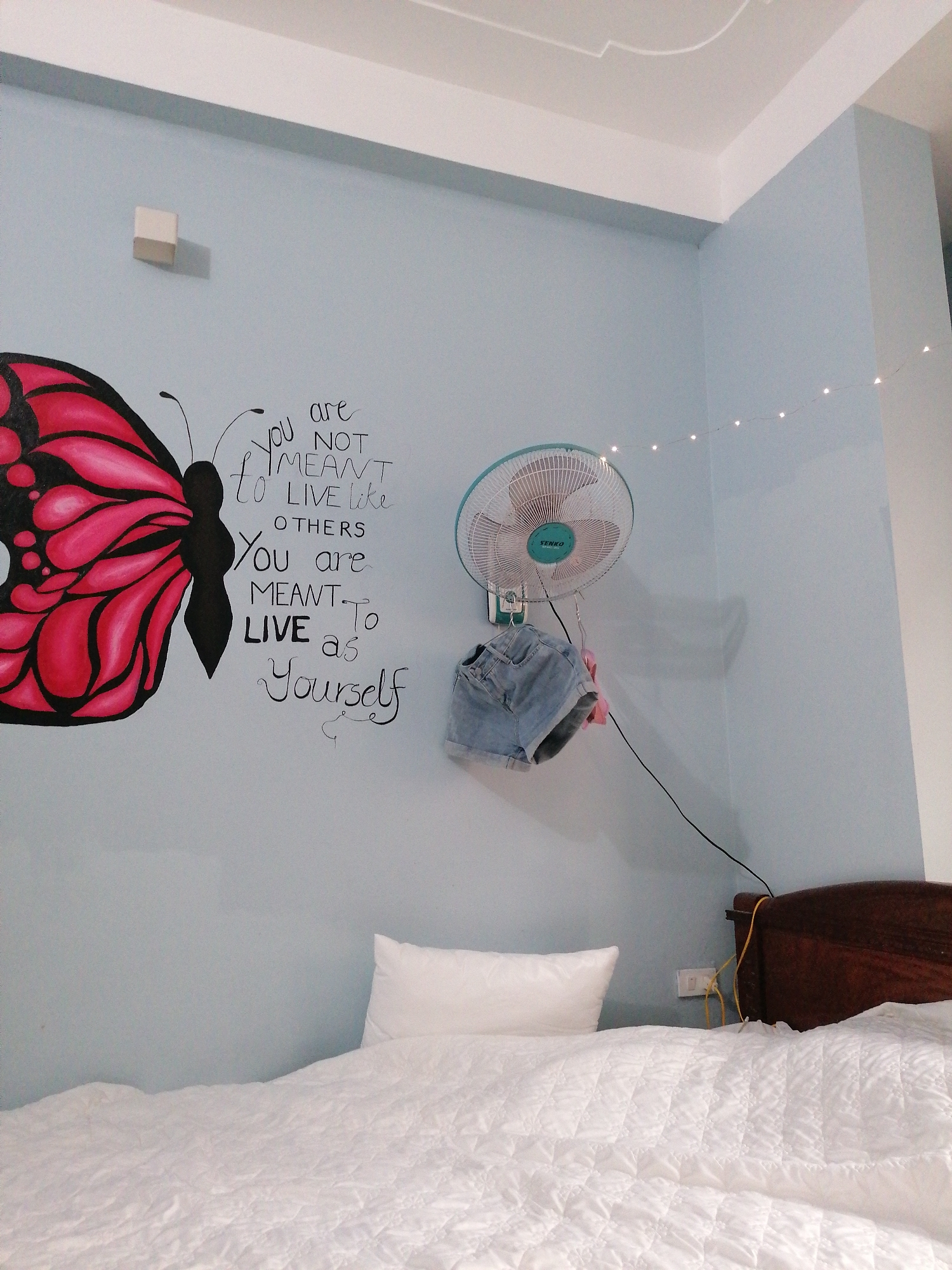 An indoor shot depicts a cozy bedroom in Bai Chay Ward, Vietnam, characterized by light blue walls, a white ceiling, and white bedding. The setting appears to be illuminated by indoor lighting, possibly in the evening given the presence of decorative string lights draped across the upper right corner of the room.
The most prominent feature is a large, hand-painted mural on the left wall, showcasing a vibrant pink and black butterfly. Beside the butterfly, a motivational quote is painted in black, combining cursive and block letters: "You are NOT MEANT to LIVE like OTHERS You are MEANT TO LIVE as Yourself."
On the adjacent wall, a white wall-mounted fan with turquoise details is visible, displaying the brand name "SENKO." A pair of light-wash denim shorts hangs casually from a hook on the fan's base. An electrical cord extends downwards from the fan.
The lower half of the image is dominated by a bed, covered with a textured white quilted comforter and a single white pillow. A dark wooden headboard is partially visible on the right, behind which an electrical outlet with a yellow cable is seen on the light blue wall. No people are present, and no specific activity is taking place, but the scene suggests a lived-in, personalized space for relaxation.