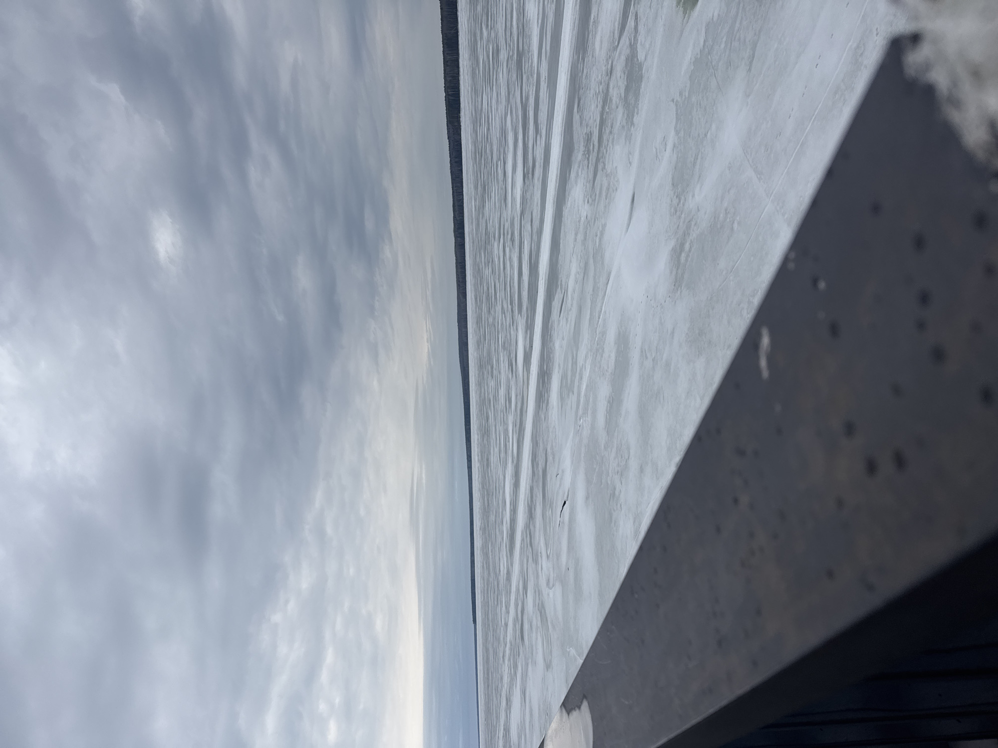 The image depicts a landscape from what appears to be a waterfront or a bridge, possibly in the city of Chaikovsky, Russia. The scene features a partly frozen body of water stretching into the distance, with a visible treeline along the opposite shore. The sky is overcast with clouds. A portion of a structure, possibly a bridge railing, is in the foreground and occupies part of the frame on the right side. The surface of the water is predominantly white and gray, likely indicating ice or snow cover. There are no visible people or animals in the frame.