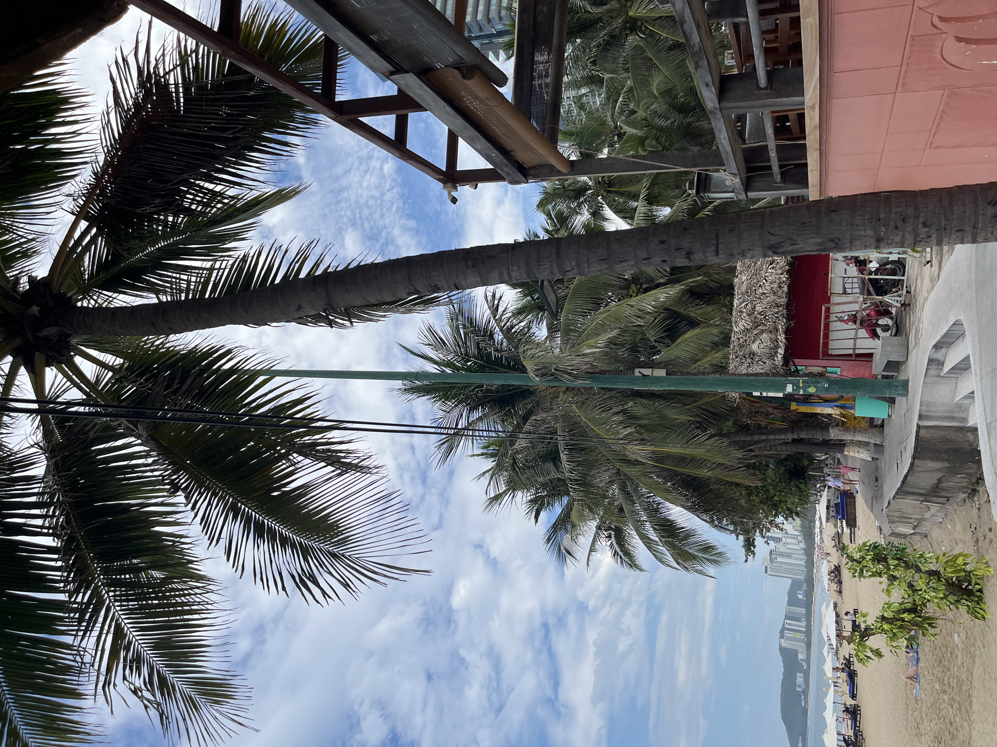 This is a low-angle, vertical, eye-level, medium shot of a beach in Nha Trang, Vietnam. In the foreground, a large palm tree trunk stands vertically on the right side of the frame, its fronds obscuring the top portion of the sky. To the right of the palm tree is a wooden structure with chairs and tables visible underneath, suggesting an outdoor seating area. A pink wall runs along the right edge of the frame, partially obscuring the wooden structure.
Behind the palm tree, a row of palm trees lines the beach. A small red building with a thatched roof is visible amongst the trees, with a red motorcycle parked in front of it. Further back, a beach with white umbrellas and lounge chairs is set up for visitors. People are scattered across the beach, some sitting under umbrellas and others walking along the shore. In the distance, modern high-rise buildings are visible on the coastline, indicating an urban environment. The sky is cloudy with patches of blue. A green lamppost is visible in the center of the frame.