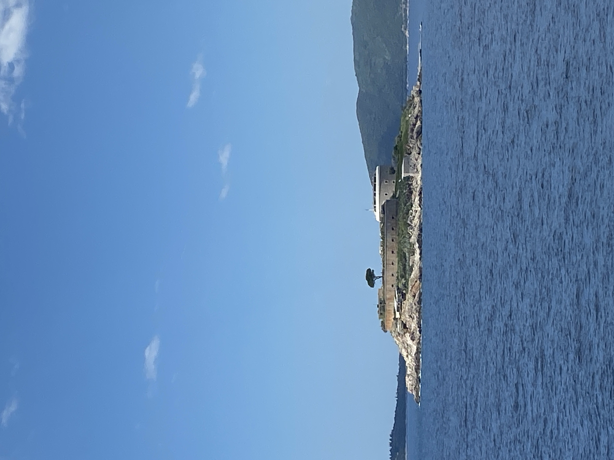 The image depicts a coastal scene in Luštica, Montenegro. A stone fortification structure is perched on a rocky cliff along the edge of a body of water, likely the Adriatic Sea. Lush green vegetation grows atop the cliff and the nearby mountain. The sky is predominantly blue with a few scattered clouds. The water surface shows the texture of small waves or ripples.