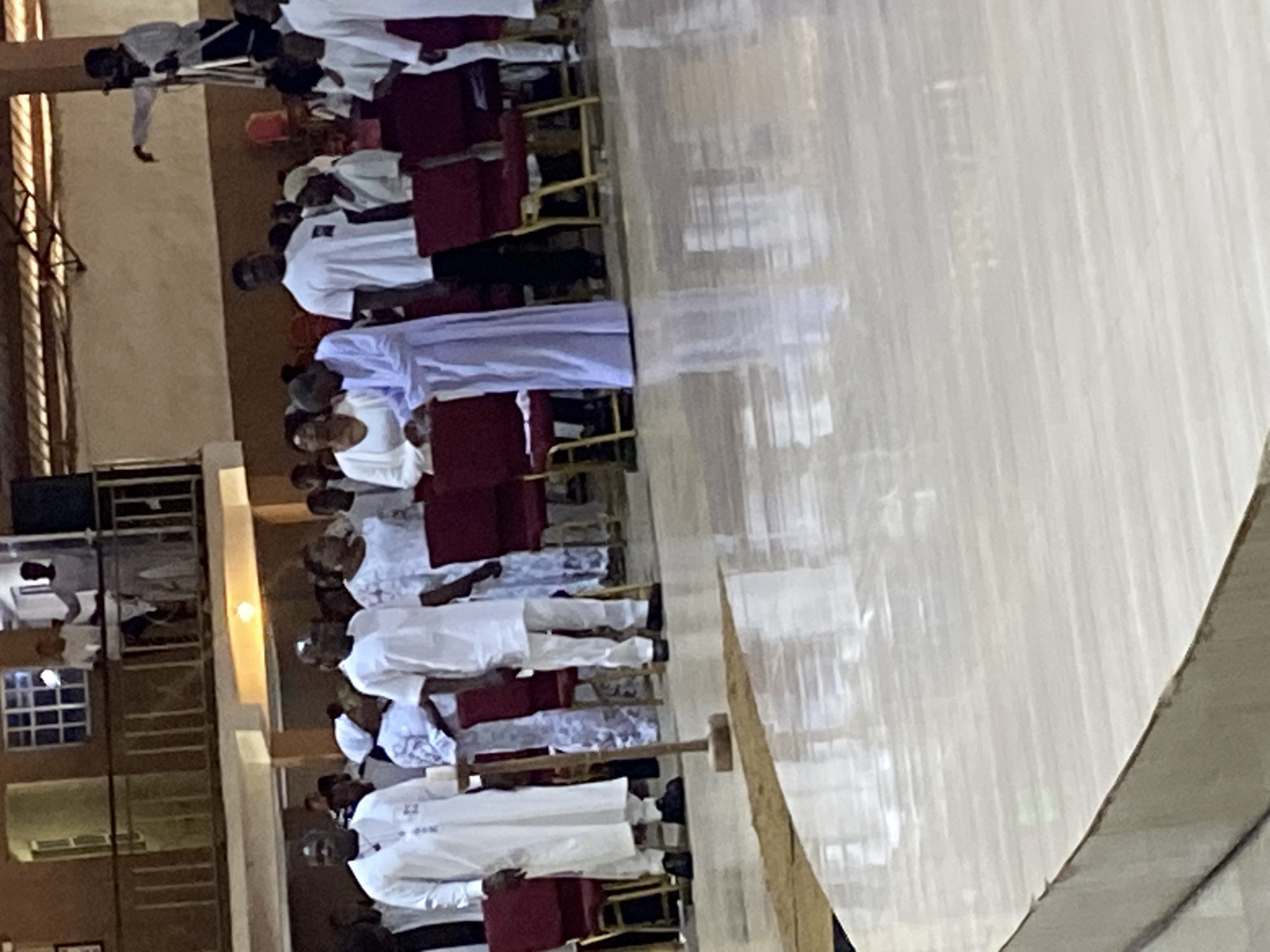 In an interior, likely a church, on Rukuba Road, Nigeria, a group of people, primarily dressed in white garments, are seated on chairs. Many of the chairs have red cushions. Most individuals appear to be adults, with variations in skin tone and age. A few are wearing white head coverings. A section of the room, potentially the aisle, is visible in the foreground, showing a shiny surface. Windows and lighting fixtures are visible in the background.