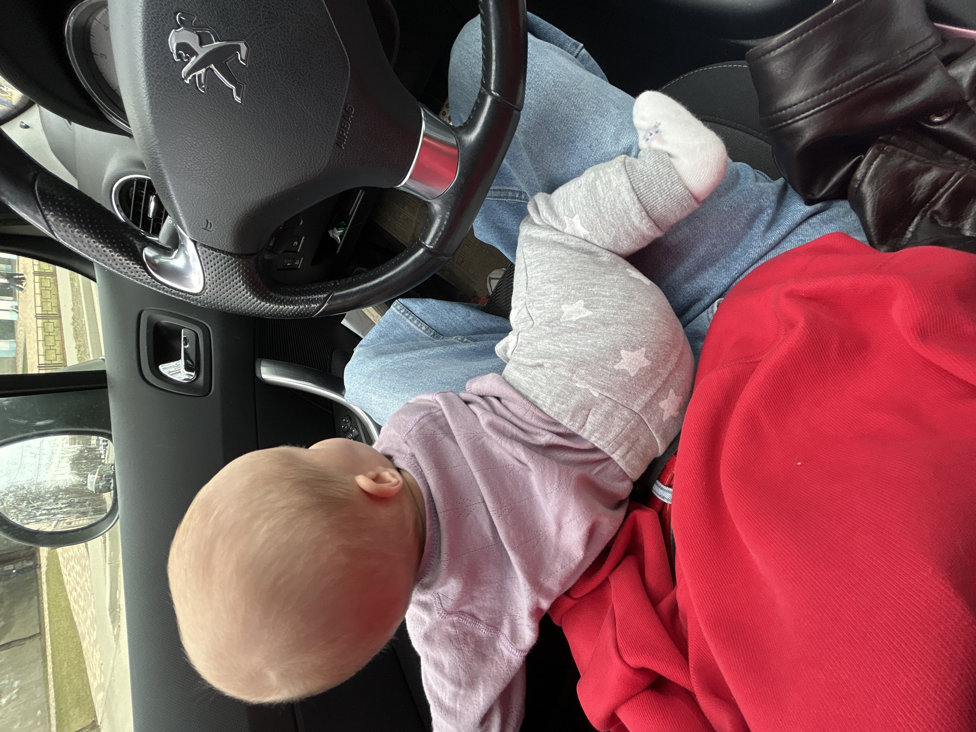 A baby lies unrestrained on the lap of a person seated in the driver's seat of a Peugeot automobile. The car interior is visible, including the steering wheel with the Peugeot logo. Outside the car window, a reflection reveals a streetscape with buildings, possibly located in Moscow, Russia based on the architecture seen. The baby is wearing gray patterned pants, a white sock, and a lavender top. A red garment and a black jacket are also in the scene.