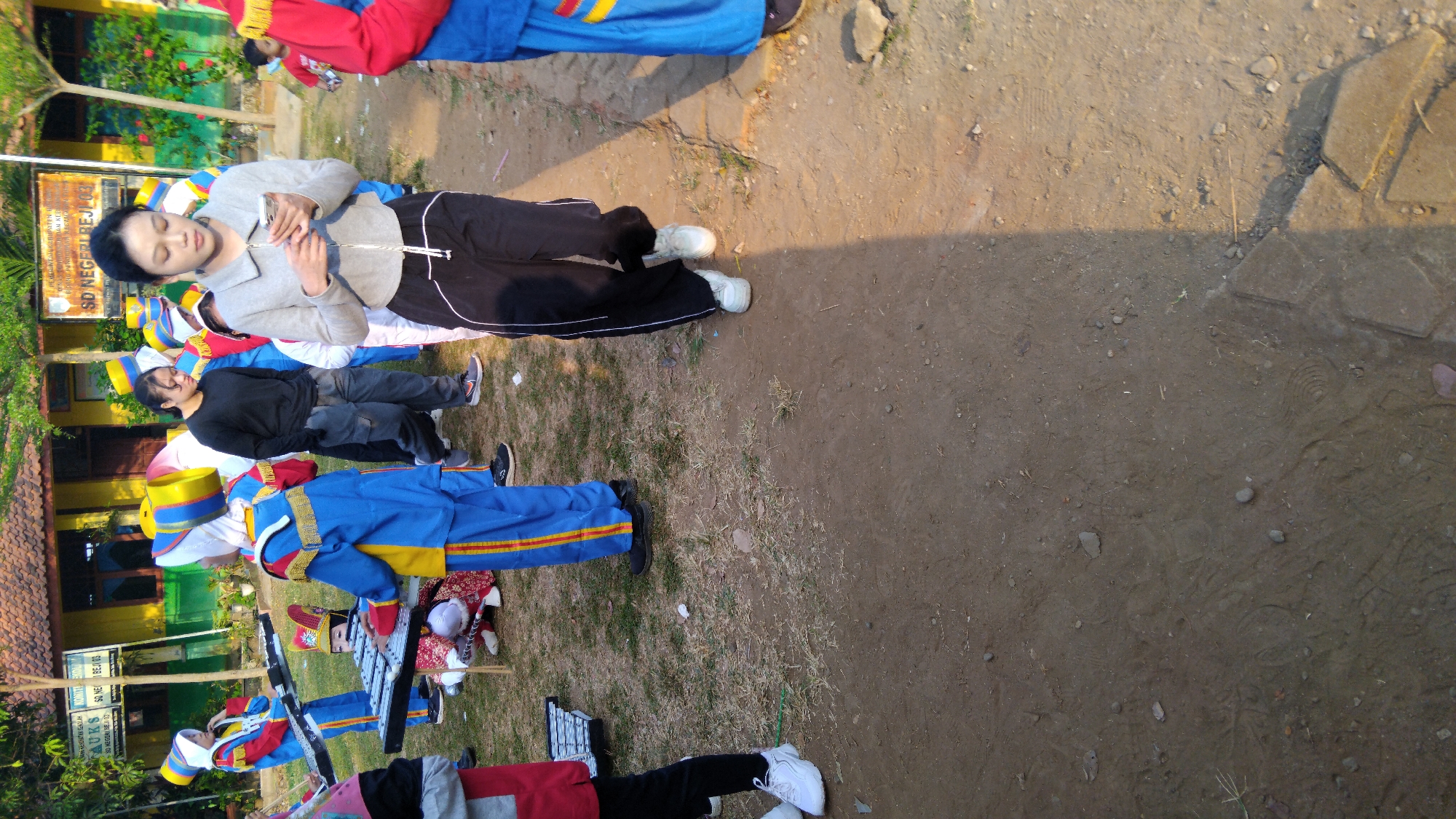 In the city of Sembojo, Indonesia, a group of individuals, some in marching band uniforms, are gathered outdoors. A young woman in a grey zip-up jacket and black pants is looking at her phone while walking. In the background, several children in blue and yellow marching band uniforms stand with instruments. Some of the children are holding what appear to be xylophones. The ground is a mix of dirt and dry grass, and a building with a yellow facade and a sign that reads "SD NEGERI REJ..." is visible. The lighting suggests it is daytime, with a strong shadow cast from the right side of the frame.