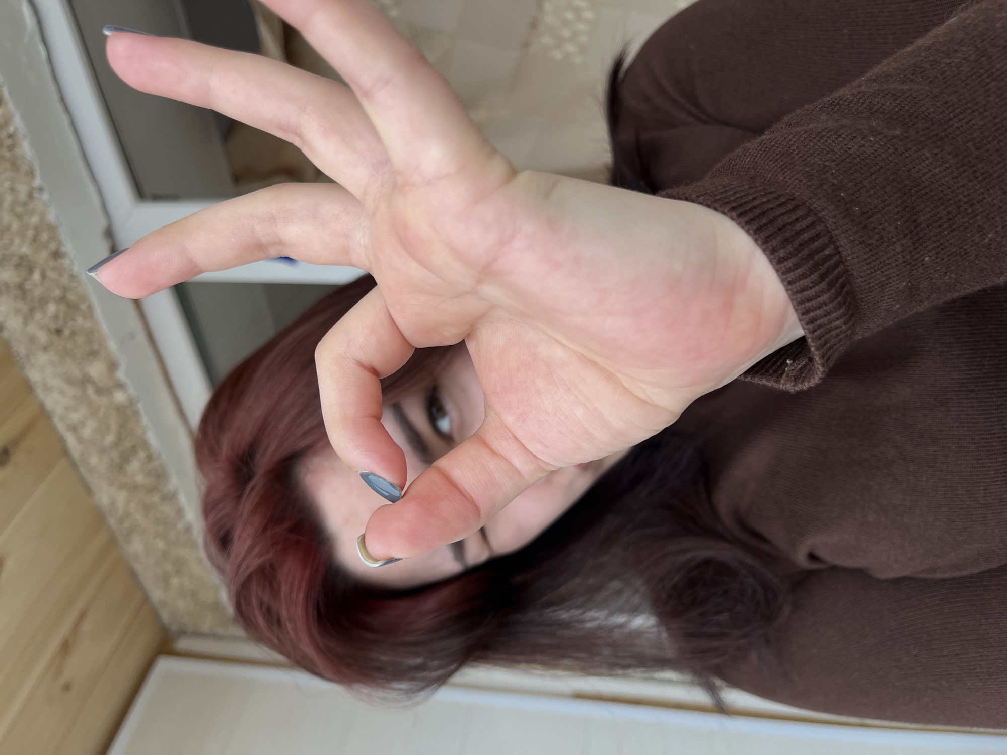 A medium close-up image shows a person, likely female, with reddish-brown hair, partially obscured by their own right hand. The person is wearing a dark brown, long-sleeved top or sweater. Their right hand is positioned centrally, forming an "OK" gesture with the thumb and index finger joined into a circle, while the middle, ring, and pinky fingers are extended upwards. The visible fingernails are painted dark grey, and the index finger displays a small, light-colored accent near the cuticle. One brown eye is visible behind the hand, appearing partially closed or winking. The background suggests an indoor or semi-enclosed outdoor area, potentially a balcony or covered porch, located in the city of Neftekamsk, Russia. Structural elements visible include a vertical white frame on the left, horizontal wooden paneling above, and a textured, light-colored wall or surface on the right. A blurred opening or window is visible further back on the right side of the background.