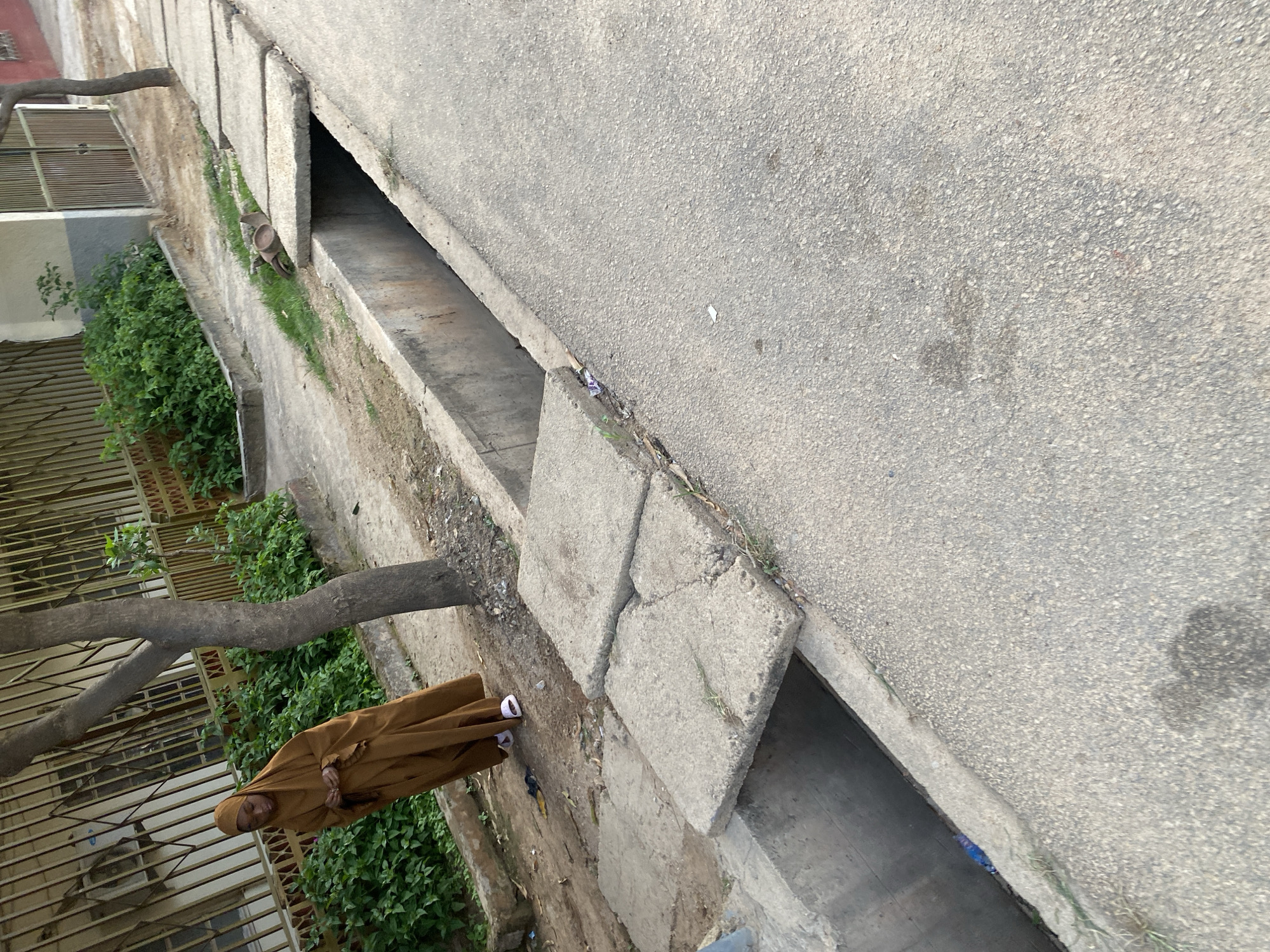 A young woman wearing a brown hijab and flowing brown dress stands with her head bowed, appearing to be praying. She is on a paved walkway next to a concrete drainage ditch. The ditch is partially covered by broken concrete slabs, and some green vegetation grows along its edges. A tree trunk is visible behind the woman. The environment appears to be an urban setting, possibly a street or sidewalk in Jos, Nigeria, with a building and a metal fence visible in the background. The ground is a mix of dirt and pavement.