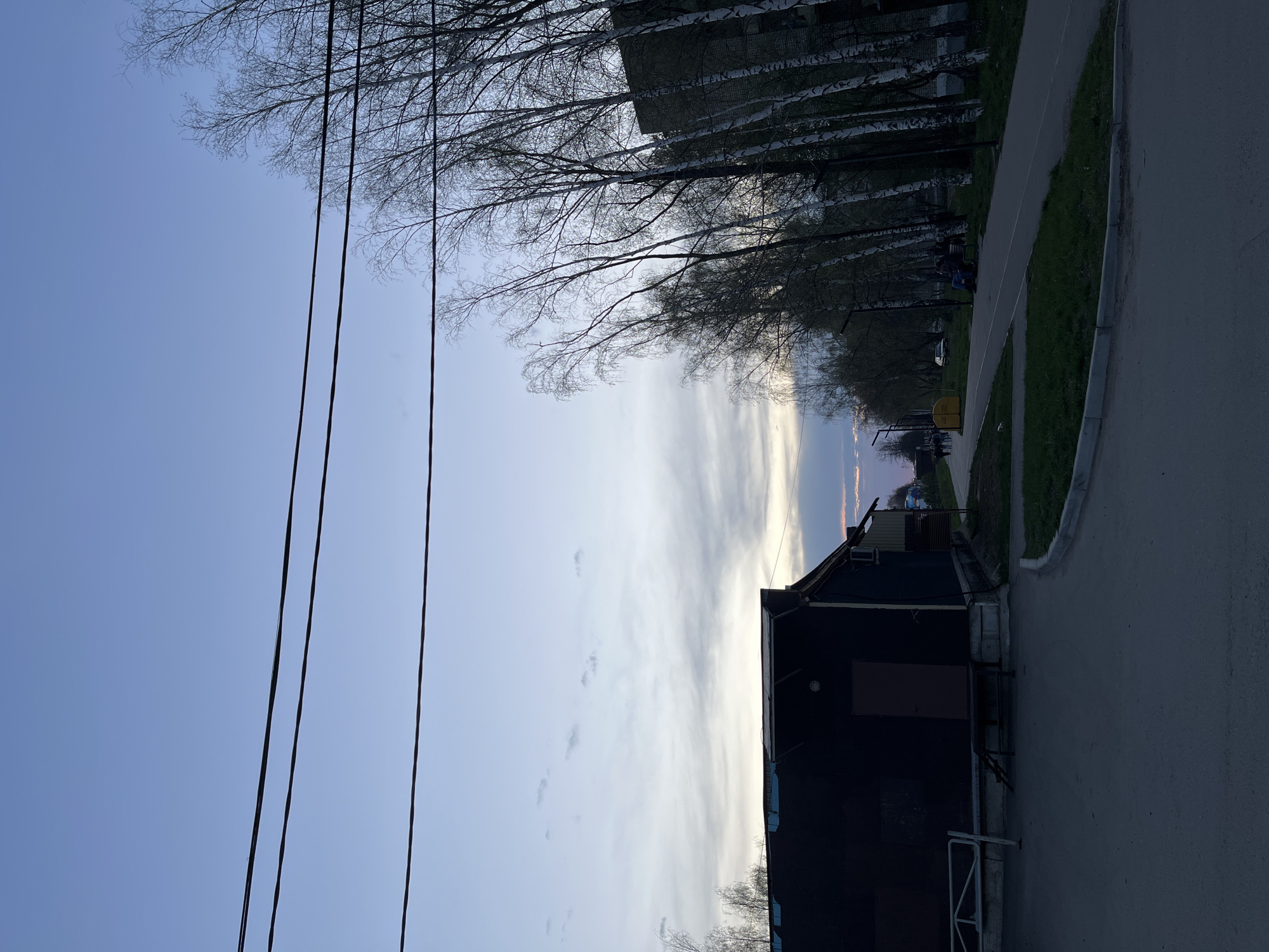 This image shows a street scene in what appears to be Chaikovsky, Russia. The perspective is from ground level, looking down a street lined with bare birch trees and a paved walkway. A building is visible on the right side of the image, and power lines are visible in the left. The sky shows a sunset or sunrise, with a blend of colors. There are people visible on a bench in the distance. The street appears to be a residential area.
