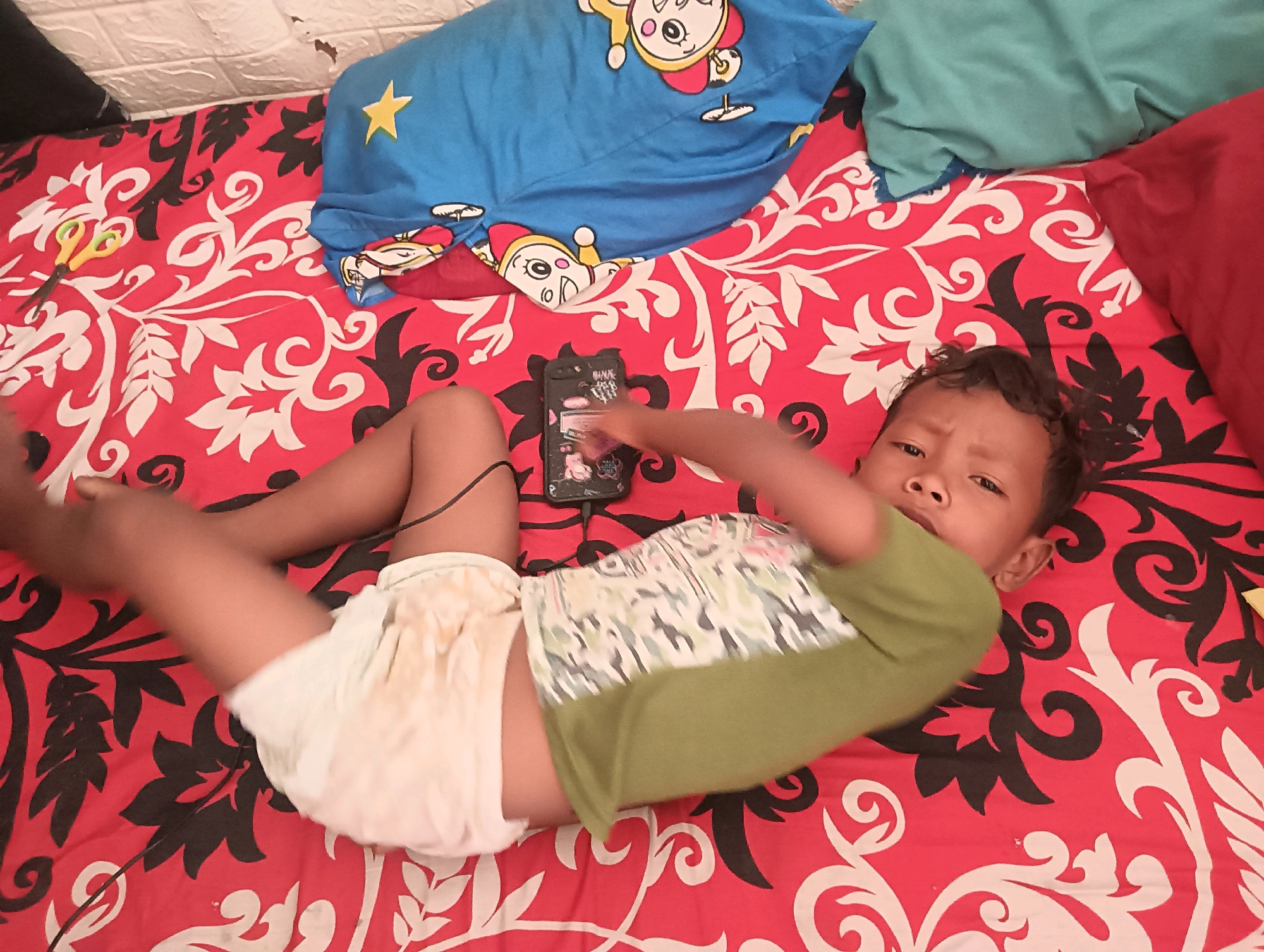 The image shows a young child lying on a bed covered with a red and white floral patterned sheet. The child is wearing a green t-shirt and a light-colored diaper. The child is looking at a mobile phone that is placed on the bed. The child's left arm is extended towards the phone, and it appears they are interacting with it. Several pillows are visible in the background, including a blue pillow with cartoon characters and a yellow star, and a turquoise pillow. The setting appears to be indoors, likely a bedroom. The time of day is unclear, but the lighting suggests it could be daytime. There is some visible text on the phone's case, which appears to be decorative stickers. The child's expression is neutral, and they seem engaged with the phone. The overall scene is casual and domestic.
