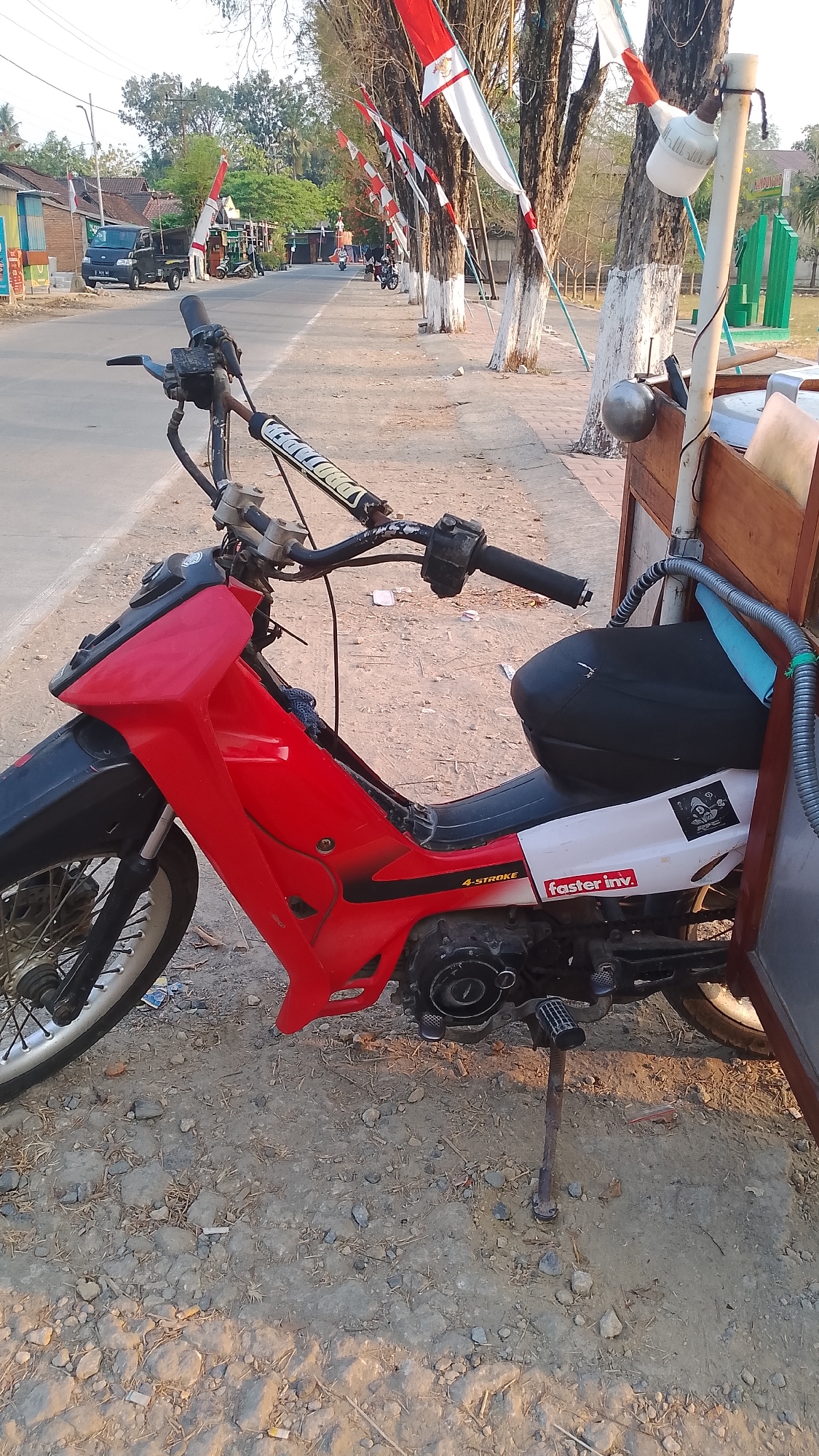 A red and black motorcycle with a sidecar is parked on a dirt and gravel roadside in Karanglangu, Indonesia. The motorcycle has a visible engine and a black seat. A wooden structure is attached to the sidecar. The road is lined with trees adorned with Indonesian flags. In the background, there are buildings, a dark-colored pickup truck, and other motorcycles. The scene appears to be in a semi-urban or rural setting.