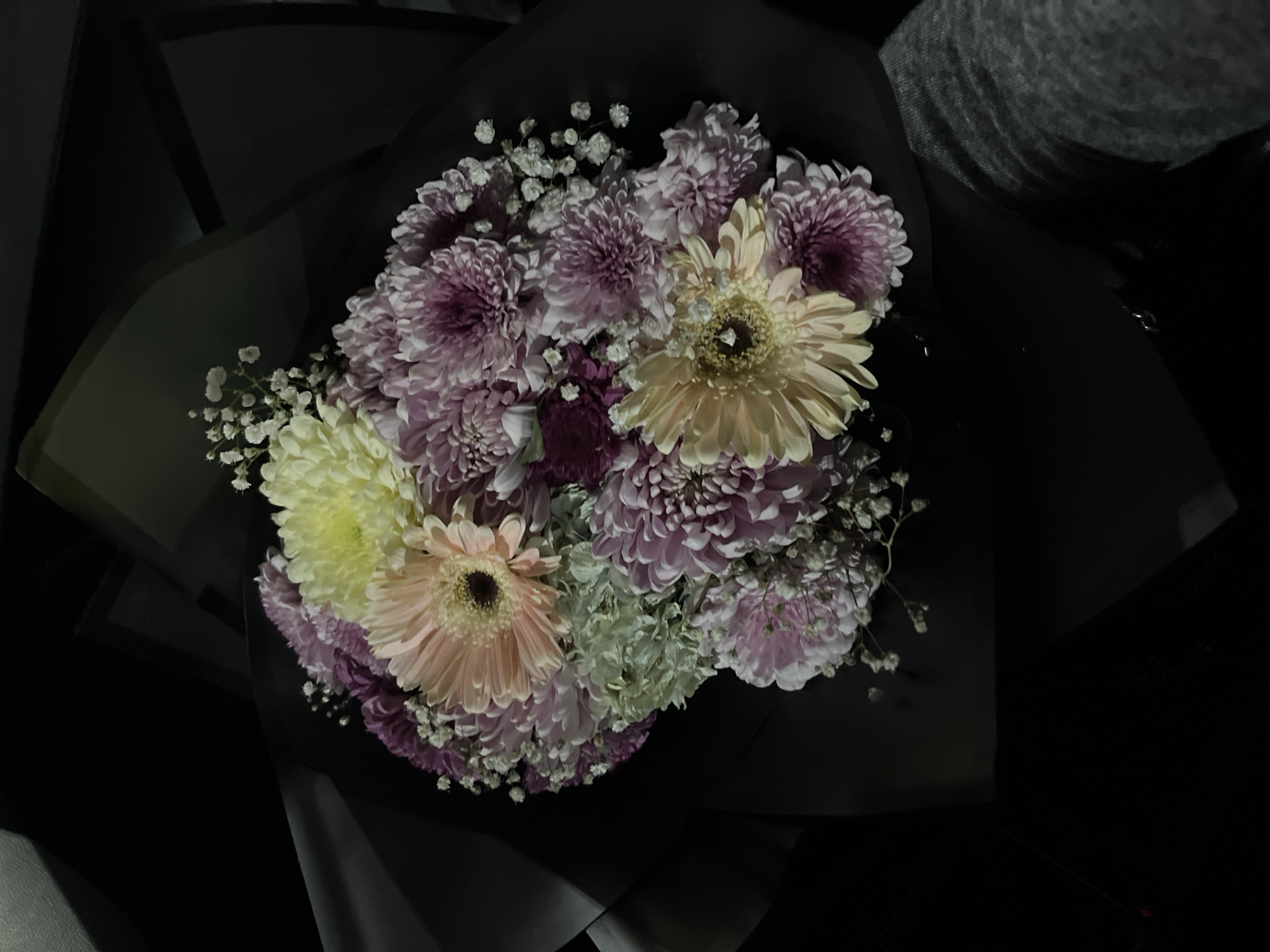 A close-up photograph displays a diverse floral bouquet, dimly lit against a dark background. The bouquet is composed of various flowers, including several prominent light pink or peach Gerbera daisies with dark centers, numerous small, dense chrysanthemum-type flowers in shades of light purple, lavender, and deeper magenta, and scattered clusters of tiny white baby's breath (Gypsophila). A single large, pale yellow chrysanthemum-type flower is visible towards the top-center. The flowers are wrapped in a dark, possibly black or very dark brown, matte paper or fabric.
The environment is poorly lit, with light originating from an unidentifiable source illuminating the bouquet primarily from above. In the lower right portion of the image, a segment of a person's leg or lap, covered in grey fabric (likely casual wear such as sweatpants), is visible, suggesting the bouquet is being held or resting in an enclosed, dark space, possibly within a vehicle or room. No other individuals or specific actions are discernible. The image provides no discernible visual information to identify a specific location in the city of Puebla, Mexico, or any other geographic area.