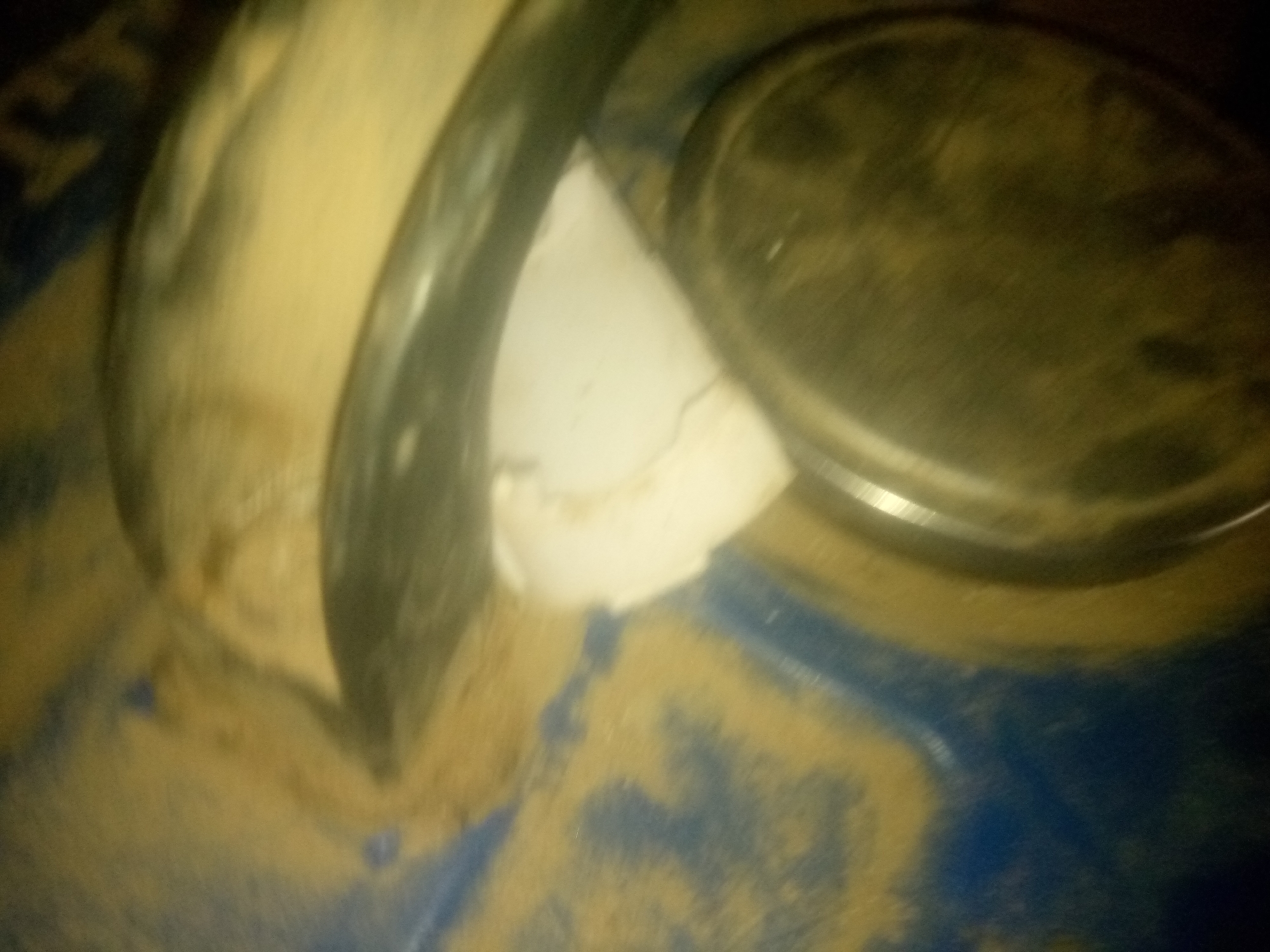 The image shows a close-up, blurry view of some objects, likely in an indoor or outdoor setting. The subjects appear to be some sort of cylindrical objects, possibly pipes or containers, covered in a light-colored dust or sand. One object is light-colored and curved, while another is darker and appears to be a lid or cap. The background is predominantly blue and brown, suggesting a potentially industrial or outdoor environment. The lighting is dim, with a yellowish cast. There is no visible text in the image. The presence of dust or sand could indicate a dry environment or recent work involving such materials. The overall impression is one of clutter or unfinished business, with the objects partially obscured by the dust. The scene does not clearly depict any specific activity or event, but rather a static arrangement of objects. The location context of Jauro Yinu, Nigeria, might suggest an outdoor setting in a region that experiences dusty conditions.