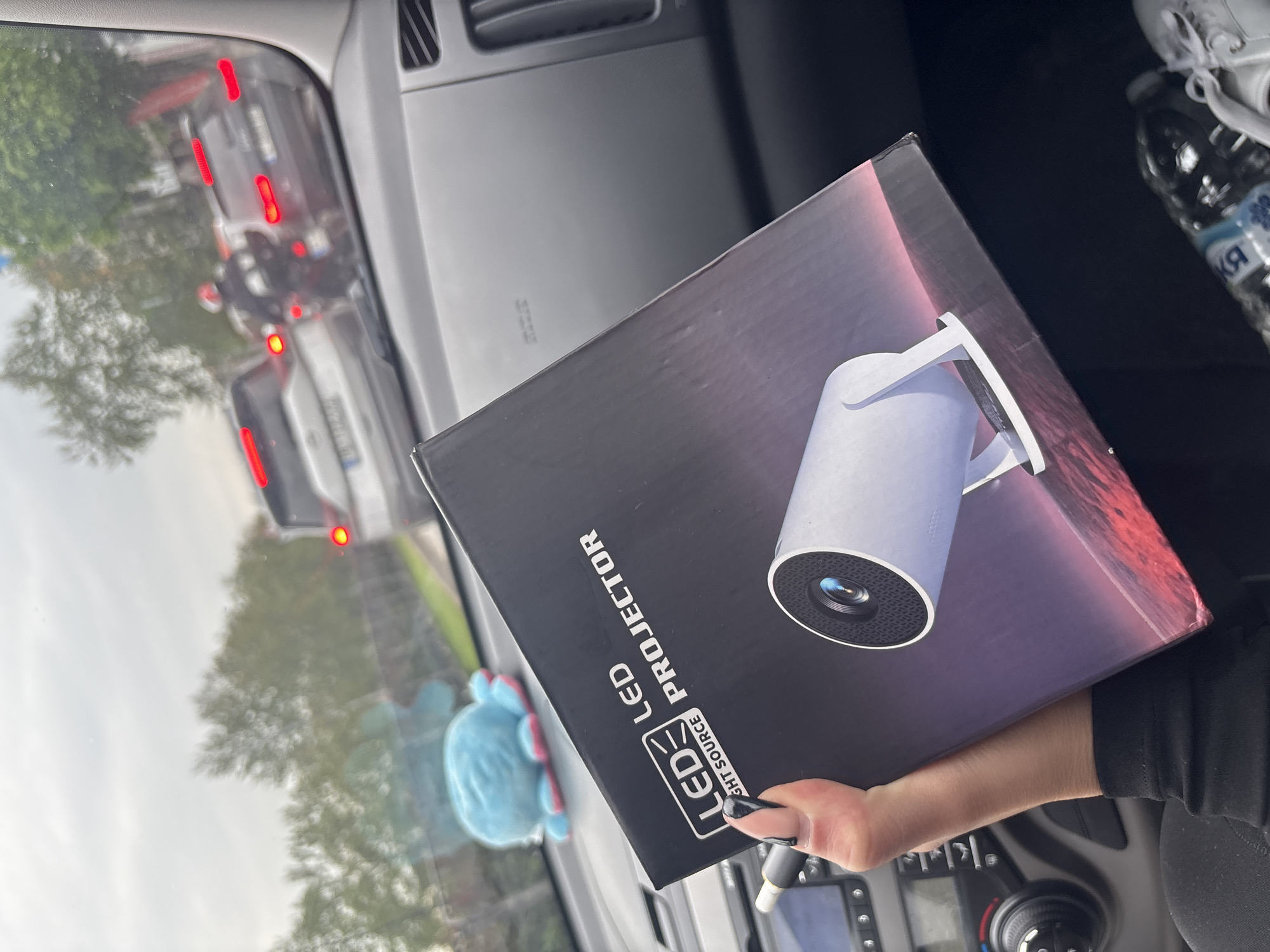 The image captures a first-person perspective from inside a car in Plovdiv, Bulgaria, with a focus on a person's hand holding a product box. The main subject is a black, rectangular box for an "LED PROJECTOR," prominently displaying the product and brand text. A white cylindrical projector is pictured on the box, with additional text indicating "LED LIGHT SOURCE." The hand holding the box has dark nail polish on the thumb.
The setting is the interior of a vehicle, with parts of the dashboard, air vents, and car controls visible. On the passenger side, a white sneaker and a plastic water bottle with Cyrillic text (likely "БЯЛА" or a similar brand) are present. Through the car window, an outdoor scene is visible, suggesting daytime with possibly overcast skies, as light is diffuse. There are reflections of trees, and a view of traffic with several cars showing illuminated red taillights, indicating slow movement or a stop. A light blue plush toy is also visible through the window. The overall activity depicts someone holding a newly acquired projector box while likely in traffic.