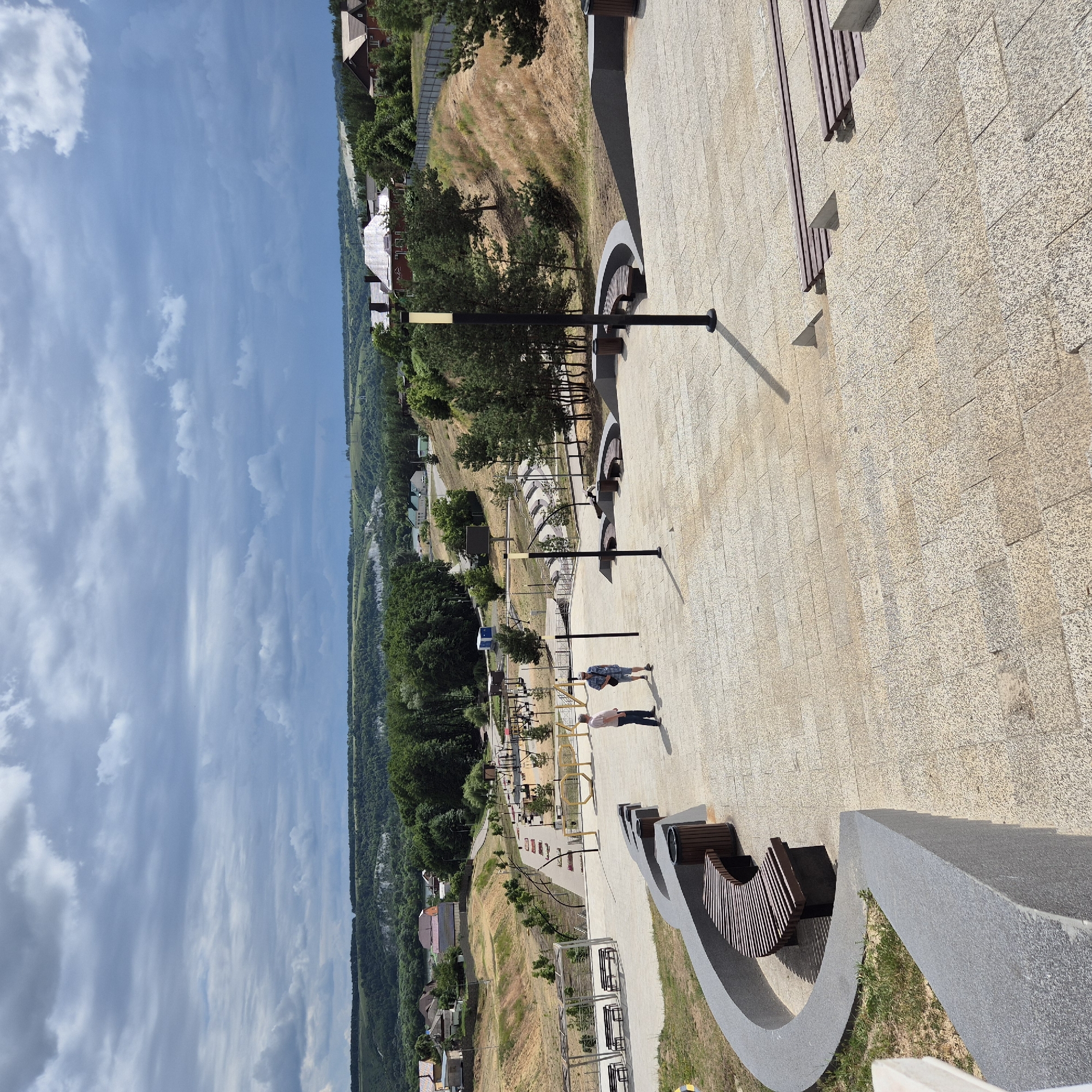 This is a high-angle, full shot image of a public park area in Liski, Russia. The park is situated on a hillside overlooking a valley with a town in the distance. The foreground is dominated by a paved area with stone steps and curved stone benches. Two men are standing on the paved area, one in a pink shirt and dark pants, the other in a plaid shirt and shorts. In the middle ground, there is a playground with various equipment, pathways, and trees. The background shows rolling hills covered in trees and houses. The sky is overcast with dramatic clouds.