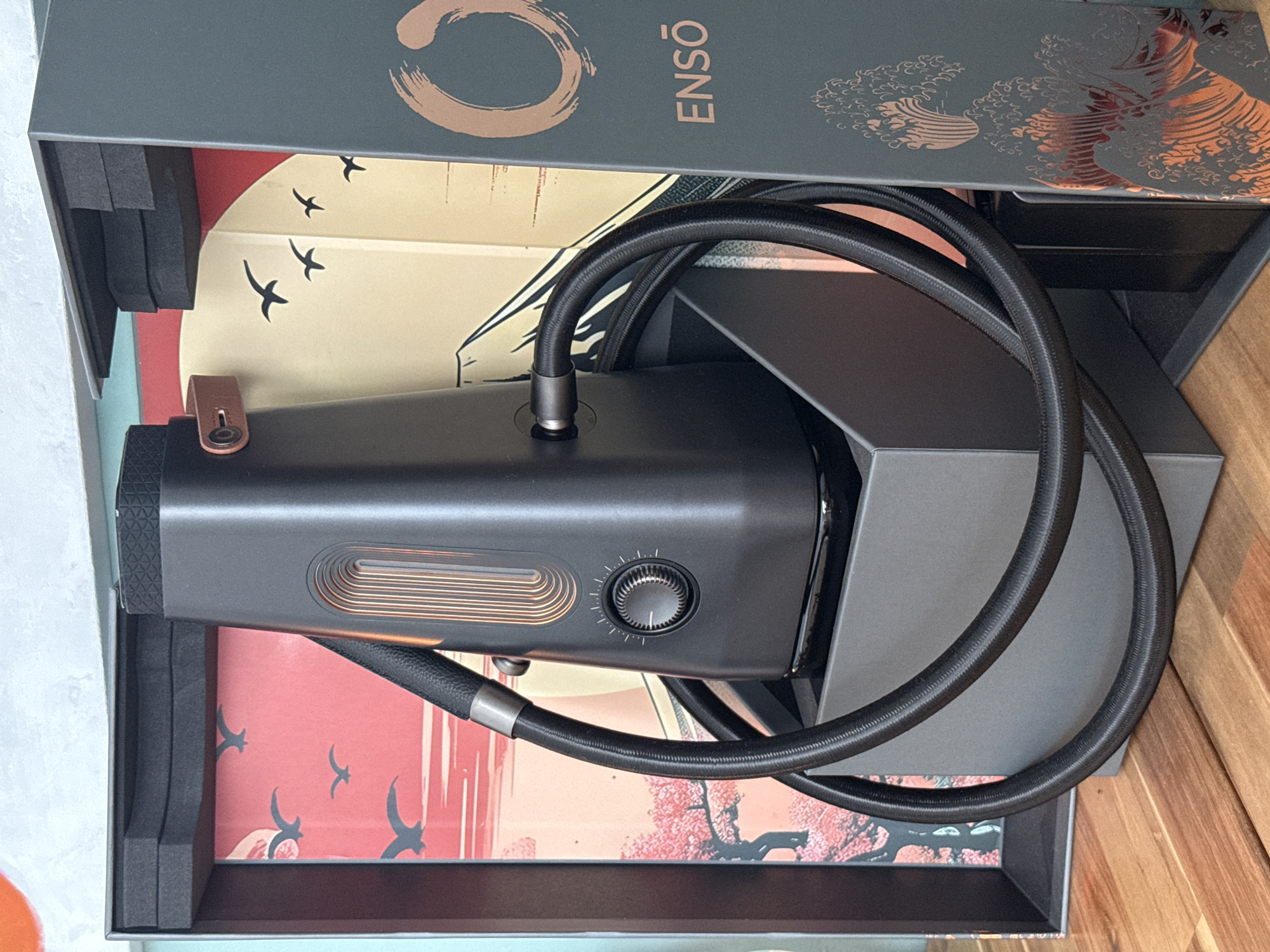 The image shows a close-up, eye-level, outdoor shot of a black electronic device with copper accents and a black hose, presented within an open box that features Japanese-inspired artwork. The device, which resembles a portable vaporizer or diffuser, has a textured top, a vertical illuminated window with copper-colored ribs, and a rotary dial with indicator markings. A flexible black hose connects to the side of the device. The box is dark gray and contains stylized imagery of birds and a mountain on the back panel, with a large copper-colored circle and the word "ENSŌ" printed on the right side. The overall aesthetic suggests a product display or unboxing. The scene is set on a wooden surface, likely a table, in what appears to be an indoor or sheltered outdoor setting, given the lighting and the backdrop. The location is not identifiable as Paphos Municipality, Cyprus, or any specific city.