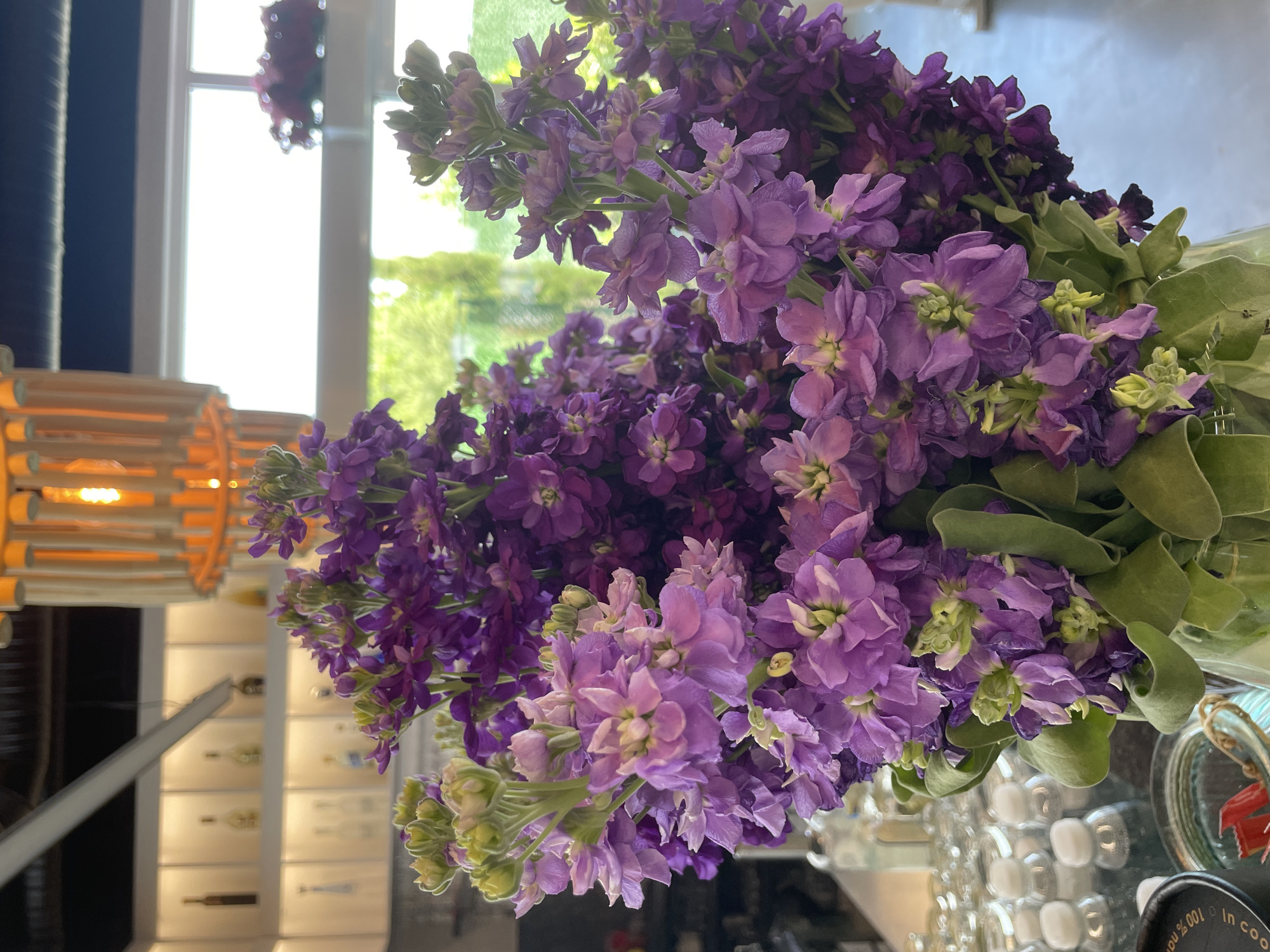 A close-up, slightly angled shot captures a vibrant bouquet of purple flowers arranged indoors. The flowers dominate the frame, their various shades of purple creating a rich, textured visual. Some green leaves are interspersed among the blossoms.
The background is blurred and suggestive of an interior space. To the left, a portion of a unique light fixture with a wooden-slat design is visible. Further in the background, a set of cabinets or drawers can be discerned, adding to the indoor setting. A window in the background provides a glimpse of natural light and what appears to be greenery outside.
The scene is well-lit, casting soft shadows and highlighting the details of the flowers. The image's focus is clearly on the floral arrangement, showcasing its colors and textures.