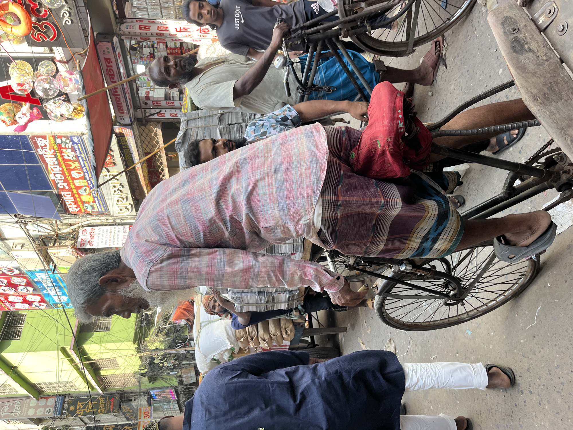 The image depicts a busy street scene in Dhaka, Bangladesh. Several men are visible, some riding bicycles loaded with goods, while others are standing or walking. The men are dressed in traditional clothing, such as lungis and kurtas, and Western-style shirts. The bicycles appear old and well-used. The background shows storefronts with signs in Bengali, along with hanging decorative items and the general bustle of a crowded urban market. The overall impression is one of a lively, densely populated commercial area.