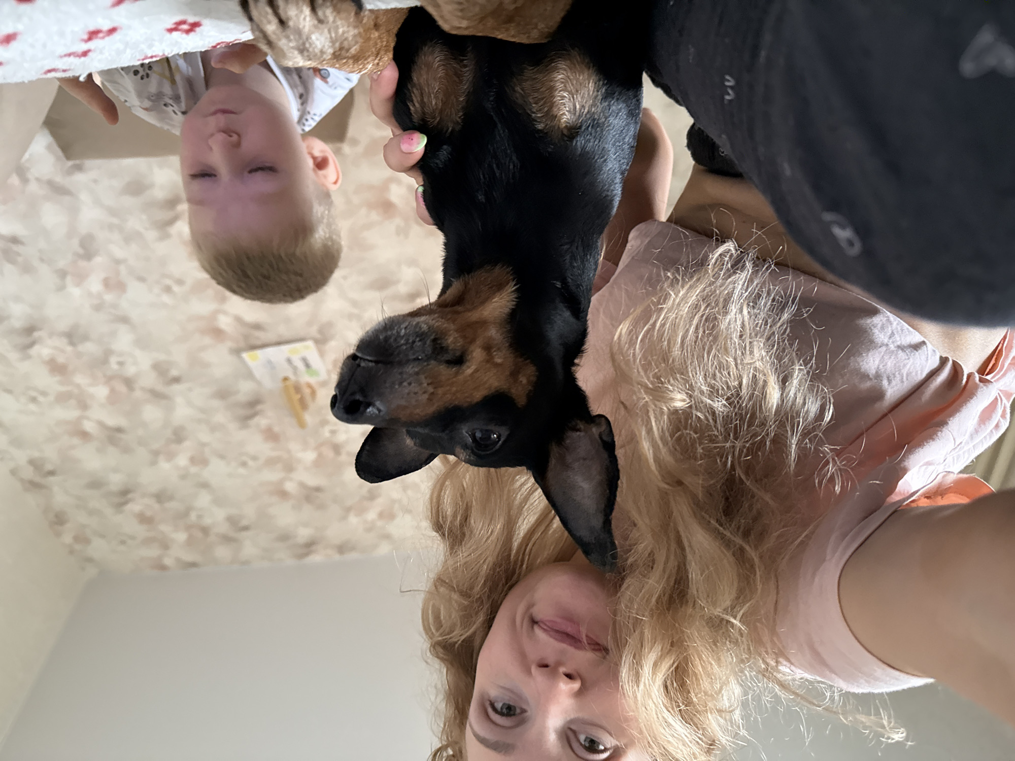 The image shows a selfie of a woman with blond curly hair, wearing a light pink shirt, holding a black and tan dachshund dog upside down. A young boy with blond hair and a white shirt is visible in the background. The background includes a wall with a floral pattern and a white wall, suggesting an indoor environment. The location cannot be determined as Perm, Russia, from the image alone.