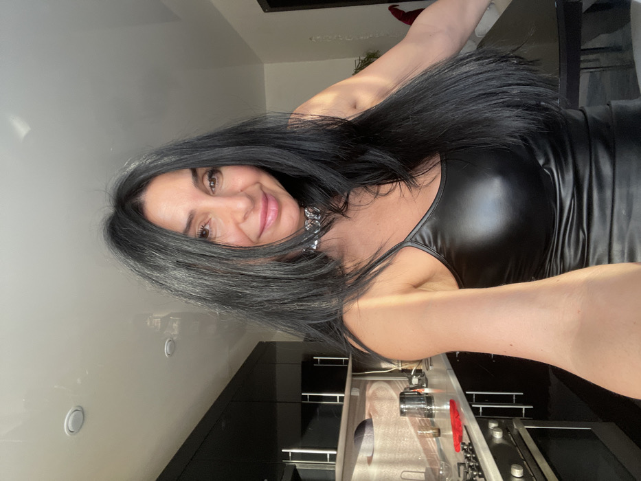 A woman with long, dark hair, wearing a shiny black top and a silver chain necklace, smiles at the camera. She stands in a modern kitchen with black cabinets and stainless steel appliances. A portion of a table or countertop is visible in the background. The image is taken at an angle, giving a tilted perspective.