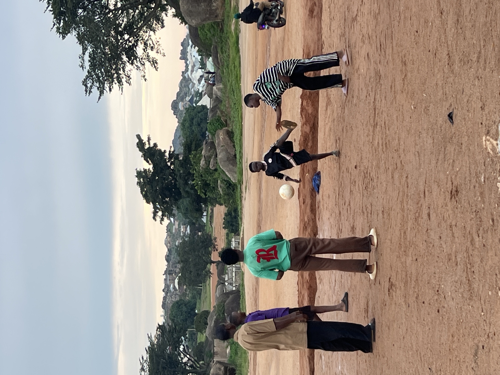In Jos, Nigeria, under a cloudy sky, a group of young men are engaged in a game of soccer on a dirt field. The field is rough and uneven, with scattered rocks and sparse vegetation in the background. In the distance, a cluster of buildings and hills are visible, suggesting a semi-urban or rural setting.
The focus of the image is the soccer game. One young man in a black and white striped shirt and black pants is about to kick a white soccer ball, while another in a black soccer jersey and shorts is positioned to receive the kick. A third young man, wearing a green t-shirt with a red "R" logo and brown pants, stands facing away from the camera, watching the game. Two other figures, one in a tan shirt and black pants and the other in a purple shirt and shorts, stand to the left, observing the match. A motorcycle is parked in the background, and a person is seated on it. The overall atmosphere appears casual and recreational.