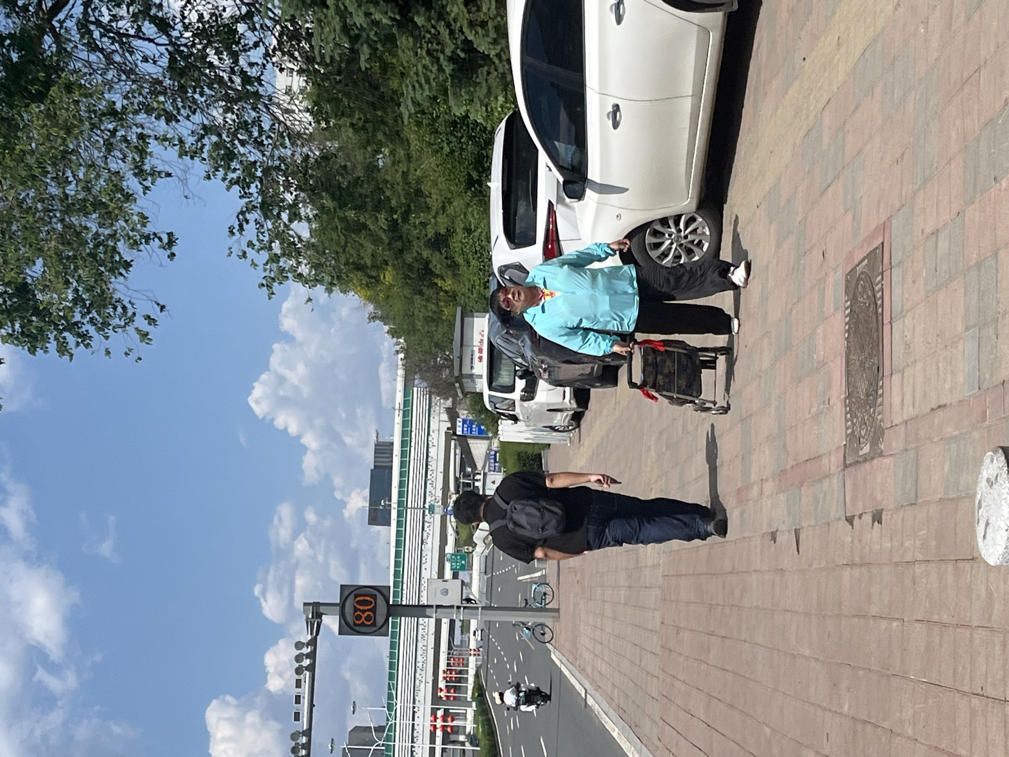 A daytime shot shows two men on a brick-paved sidewalk along a city street in Changchun City, China. One man, wearing a light blue shirt, dark pants, and white shoes, stands next to a parked white car with a cart; the man appears to be injured near his eye. Another man wearing dark clothing walks further down the sidewalk, away from the first man. The street has several cars, a bicycle, and a motorcycle traveling on it. A sign reads "80" above the street, and trees and buildings are visible in the background. The sky is blue with scattered clouds.