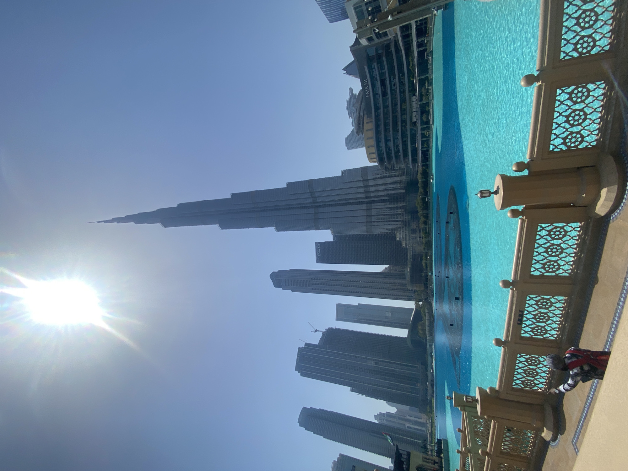 Here is a description of the image:
The image captures a daytime view in Dubai, United Arab Emirates, showcasing the Burj Khalifa, the world's tallest building. The skyscraper dominates the skyline and is partially obscured by the bright sunlight in the top left corner of the frame. Several other skyscrapers are also visible in the image, forming part of the city's modern architecture.
In the foreground is the Dubai Fountain, with light blue water in its pool. To the right of the fountain, there is an ornate fence with an Arabic design, possibly part of a viewing platform or a walkway, behind which there is a figure of a person, possibly wearing a checkered red-and-white shirt, with dark pants. The angle of the camera is pointed slightly upwards, towards the Burj Khalifa.