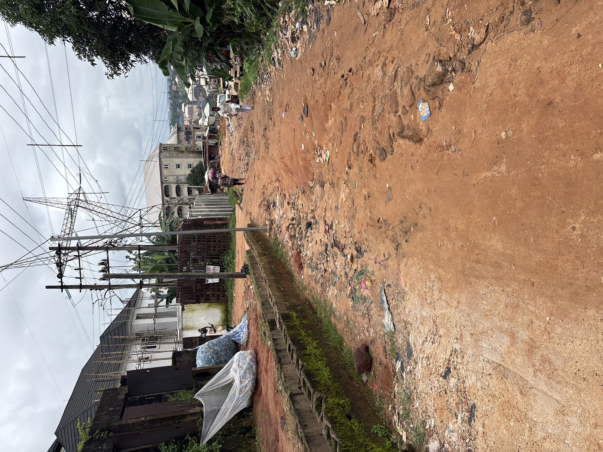 This image depicts a street in Nkpor, Nigeria, characterized by a dirt road with numerous potholes and a significant amount of debris and trash scattered along its sides and on the road itself. The environment appears to be a mix of residential and commercial areas, with buildings of varying sizes and architectural styles visible in the background. Overhead power lines crisscross the sky, supported by electrical poles. In the foreground, a concrete drainage channel runs along the side of the road, partially overgrown with moss and grass. Two people are visible walking on the road. One person, dressed in white, is walking away from the camera towards the upper right of the image. Another person, wearing darker clothing, is walking towards the lower left of the image. Several large bags, likely containing waste or recyclable materials, are placed on the side of the road near a fence. The overall impression is one of neglect and poor infrastructure.
