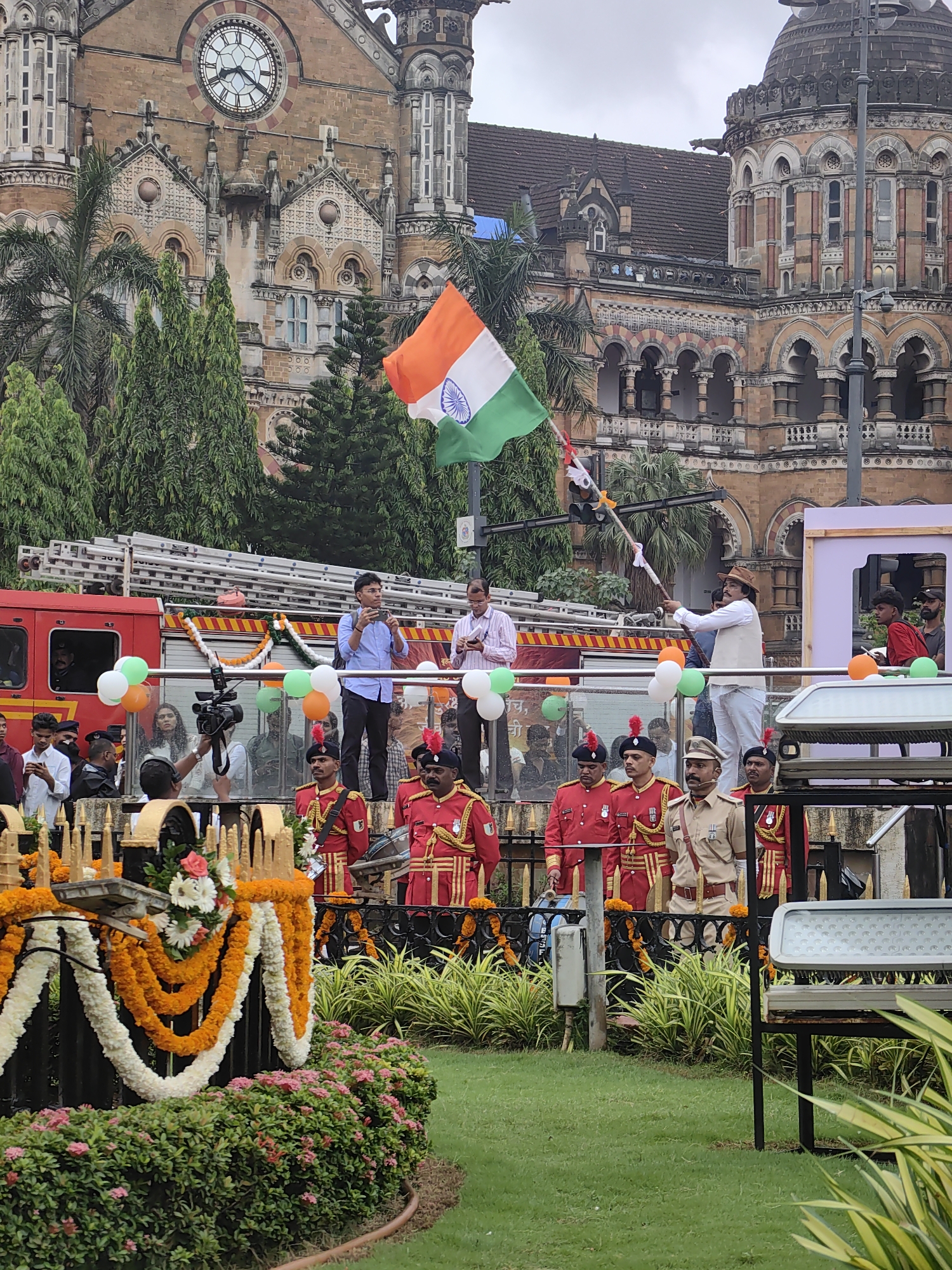 The image depicts an outdoor event, likely a celebration or parade, in front of the Victoria Terminus railway station, now known as Chhatrapati Shivaji Maharaj Terminus, in Mumbai, India. A large, ornate brick building with a clock tower and dome dominates the background. In the mid-ground, a red fire truck with a ladder is visible. Several people are standing on a platform or elevated area. Some are in ceremonial red uniforms with black hats and gold trim, while others are in civilian attire. One man in a white shirt, beige vest, and brown hat is holding a flagpole with the Indian flag. The Indian flag, with its saffron, white, and green stripes and the Ashoka Chakra in the center, is prominently displayed and waving. Green, white, and orange balloons are also visible, adding to the festive atmosphere. In the foreground, there is a green lawn with a decorative black fence adorned with orange and white flower garlands. The overall scene suggests a national celebration, possibly Independence Day or Republic Day.