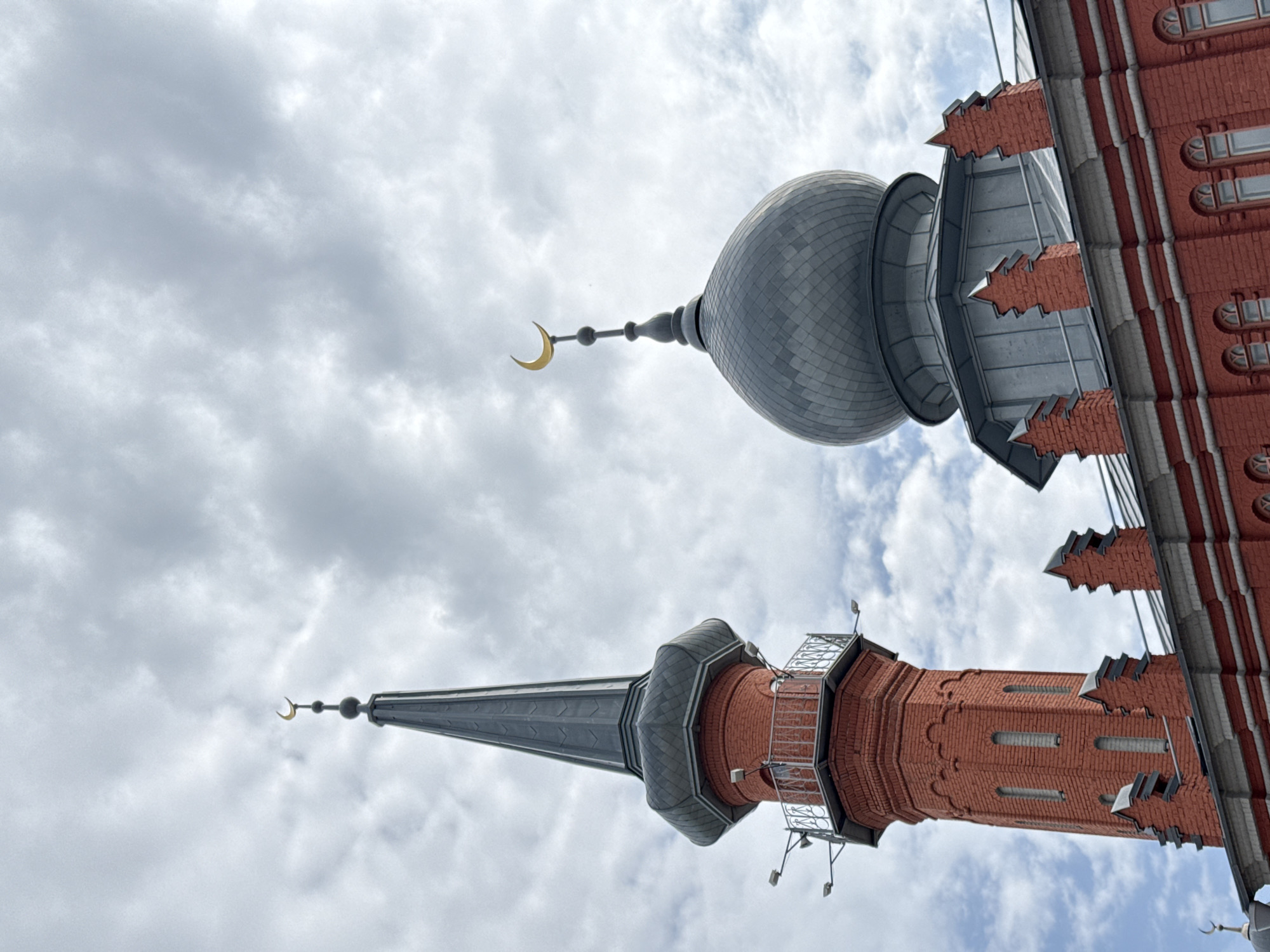 The image shows an upward view of two minarets against a cloudy sky. The minarets are part of the Nizhny Novgorod Mosque, located in the city of Nizhny Novgorod, Russia. One minaret features a gray dome with a golden crescent moon at the top. The other minaret is taller and more pointed, also with a crescent moon. Both structures are made of red brick and gray metal. The sky is filled with clouds, suggesting overcast weather.