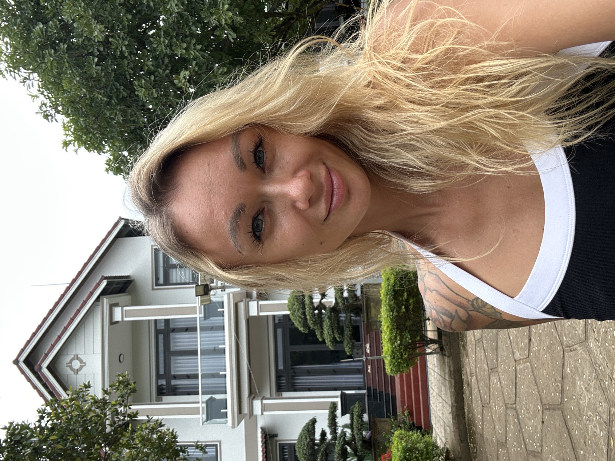 A blonde woman with blue eyes is standing in front of a two-story house in Thôn 2, Vietnam. The house is white with a red tiled roof and has a balcony on the second floor. There are trees and bushes around the house, and the ground is paved with stone. The woman is wearing a black and white tank top and has tattoos on her arm. She is smiling and looking at the camera. The sky is overcast.