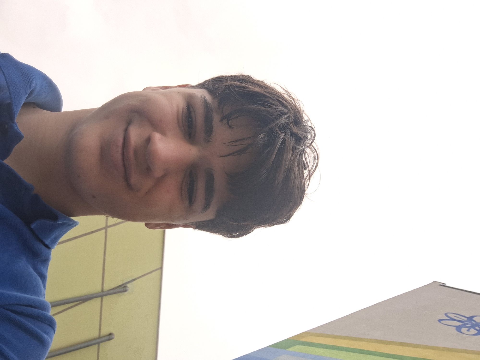 A selfie taken from a low angle of a young man with brown hair, wearing a blue polo shirt, is partially visible. He is smiling softly. The background includes a section of a yellow building with a grid-like pattern and metal supports. Another structure, possibly a sign or building facade, with bands of blue, green, and yellow paint is also visible. In the top right corner, the sky is bright white and featureless. Based on the presence of the buildings and the prompt, the photo was likely taken in Burgas, Bulgaria.